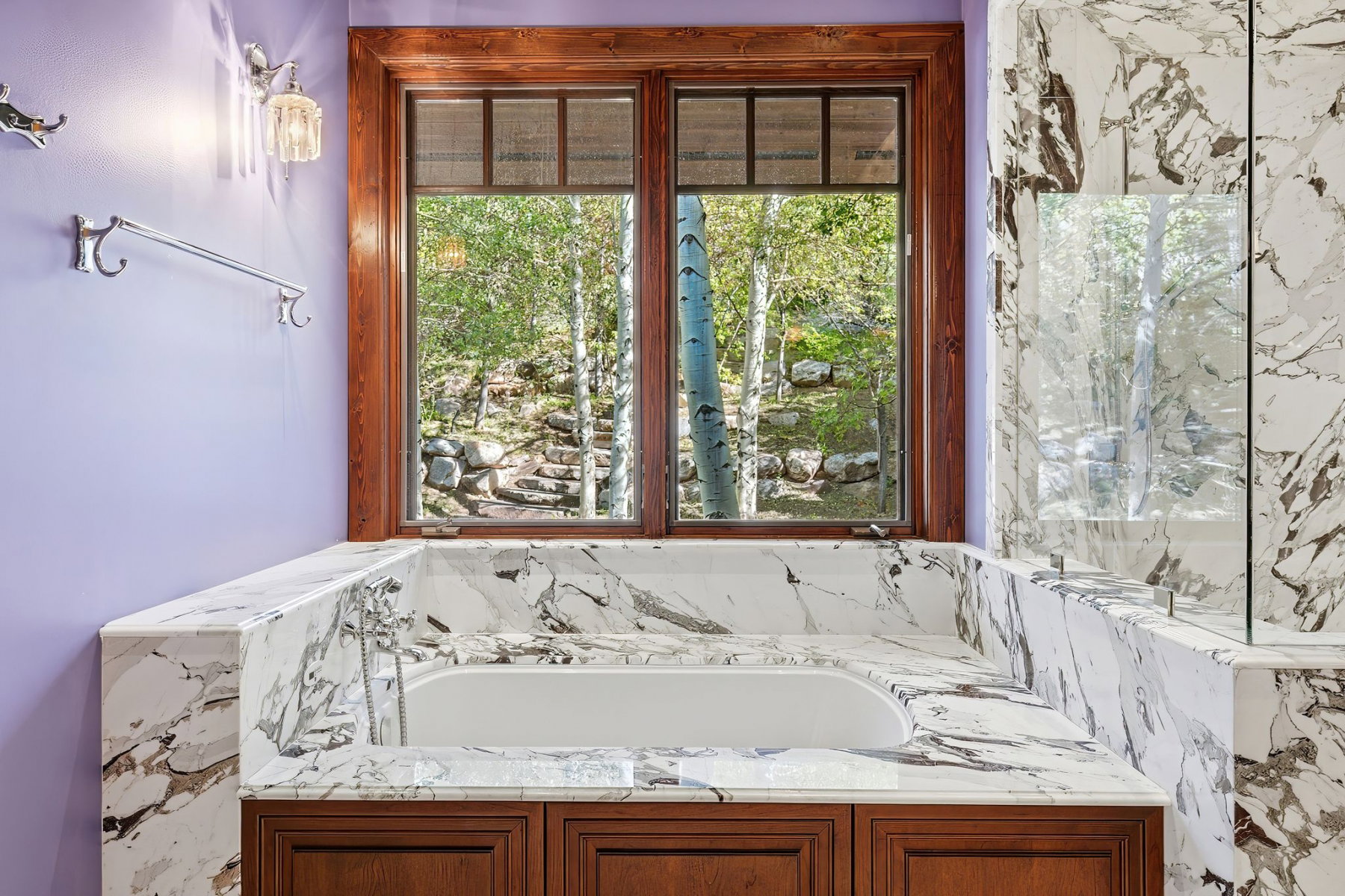 Built-in soaking tub with extensive marble surround, wood-frame window overlooking natural scenery, integrated shower partition, and wood-finish cabinetry