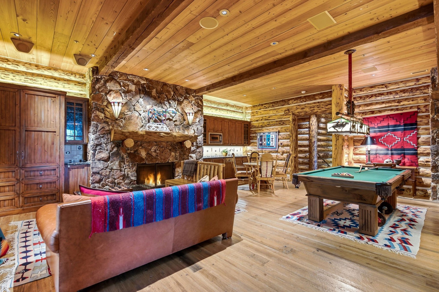 Spacious great room featuring a floor-to-ceiling stone fireplace with a substantial timber mantel, wood-finish flooring, exposed timber ceiling beams, and log-style wall construction