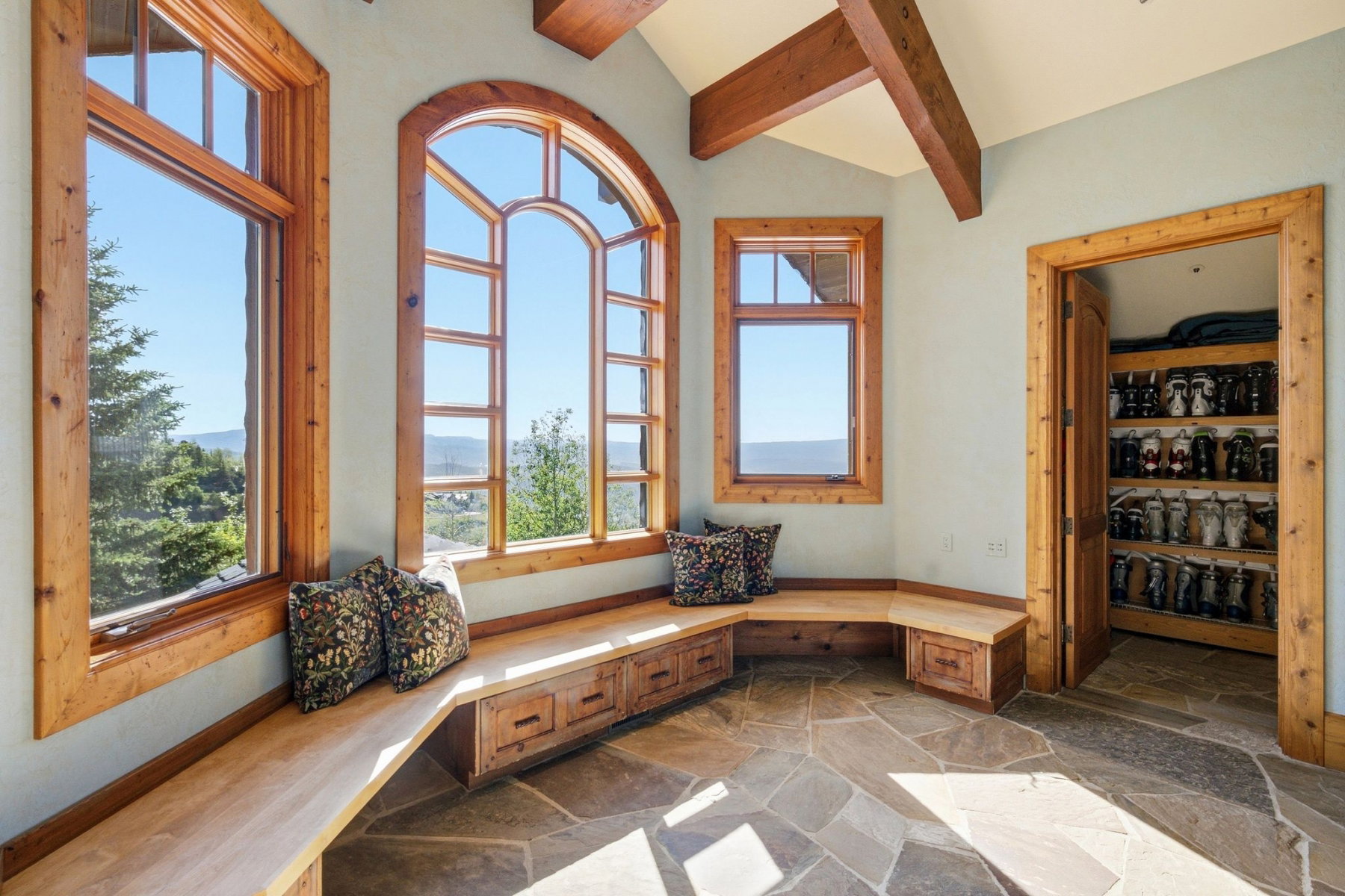 Spacious sunroom featuring an arched window, multiple rectangular windows, built-in window seating with storage, exposed ceiling beams, and flagstone flooring