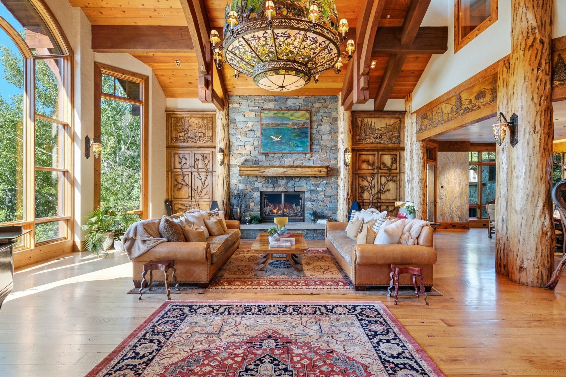 Spacious living area featuring a stone fireplace with a substantial wood mantel, exposed ceiling beams, wood-finish flooring, and expansive windows