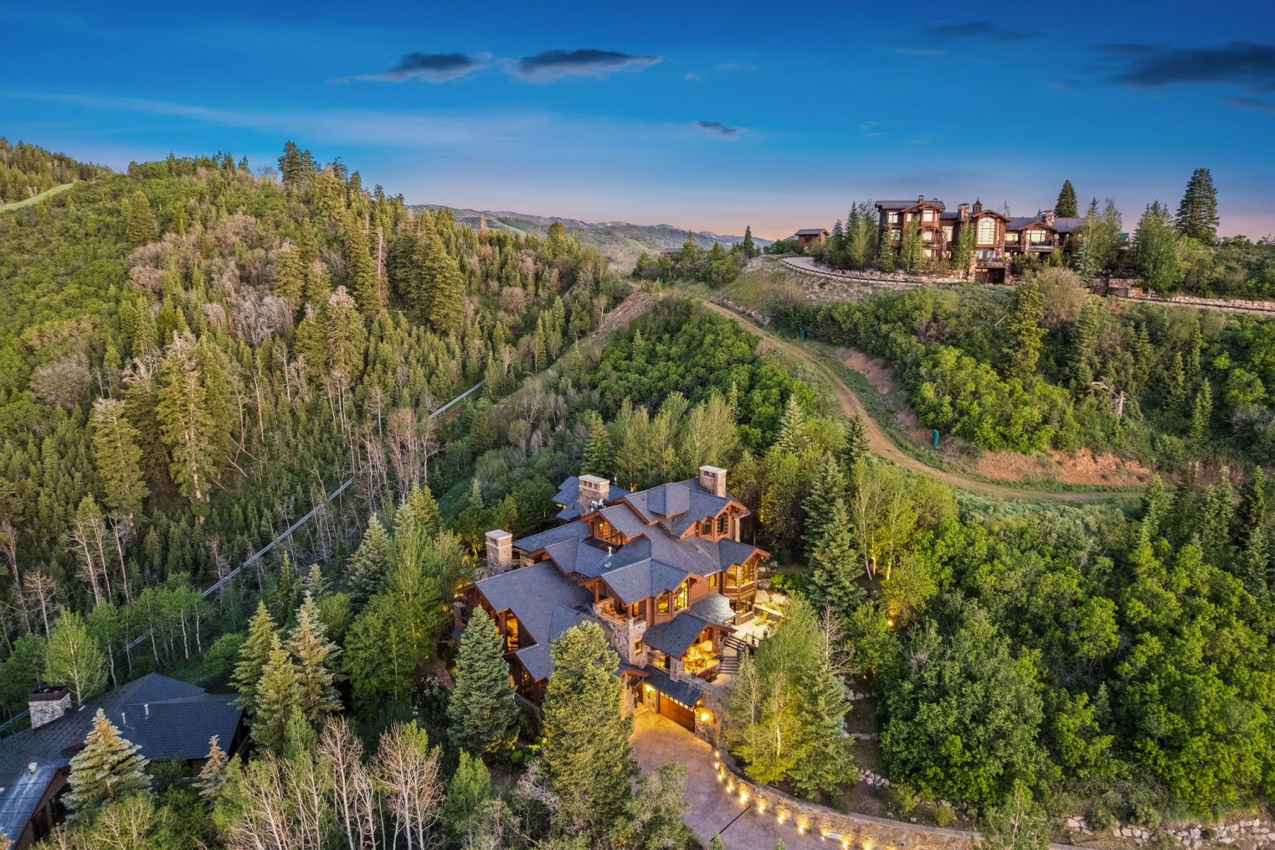 Hillside property featuring extensive tree coverage, multiple stone chimneys, a dark shingle roof, and a paved driveway with integrated lighting