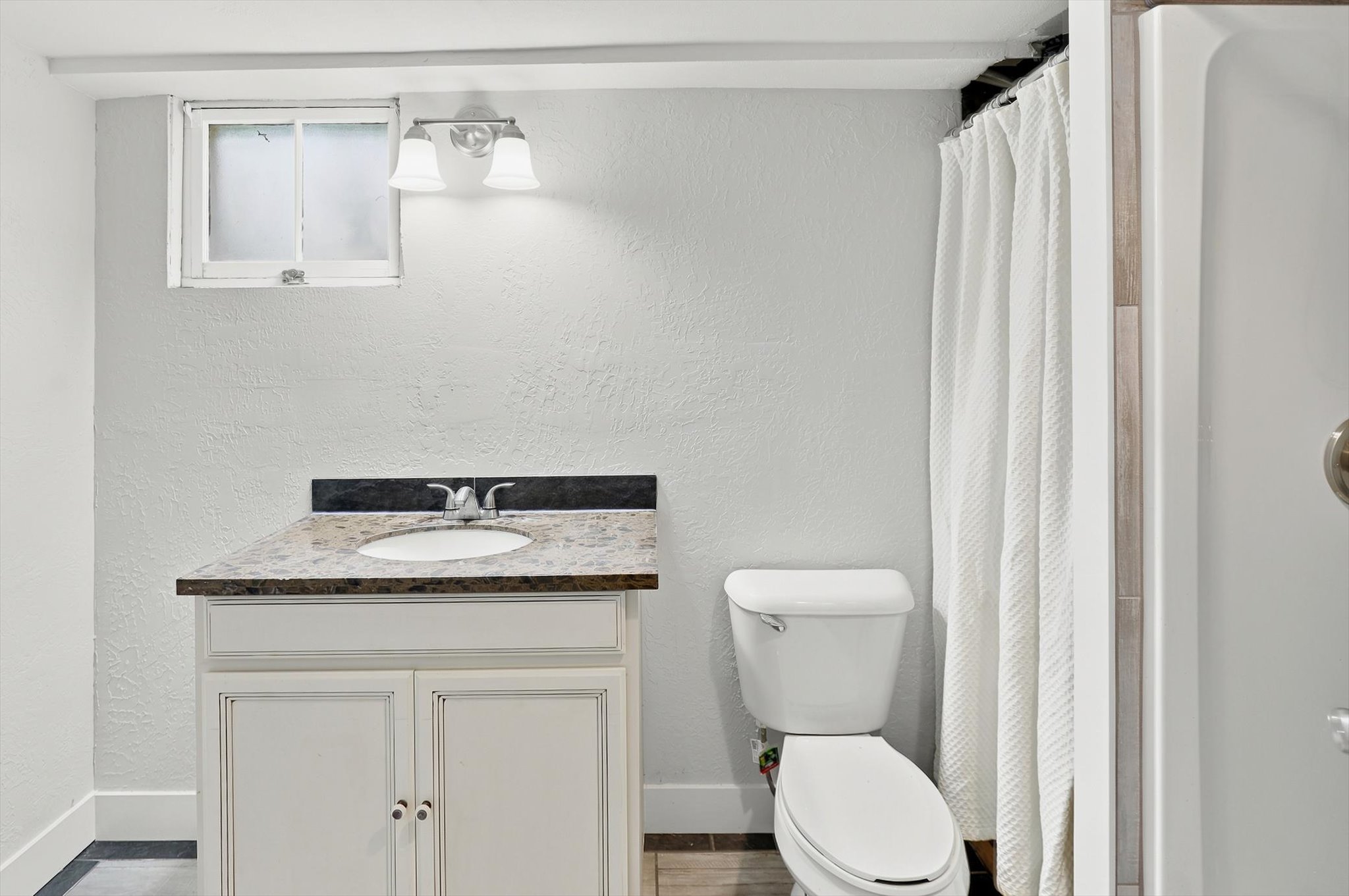 Bathroom featuring a single-sink vanity with a speckled countertop, a toilet, a privacy window, and an enclosed shower with a white shower curtain