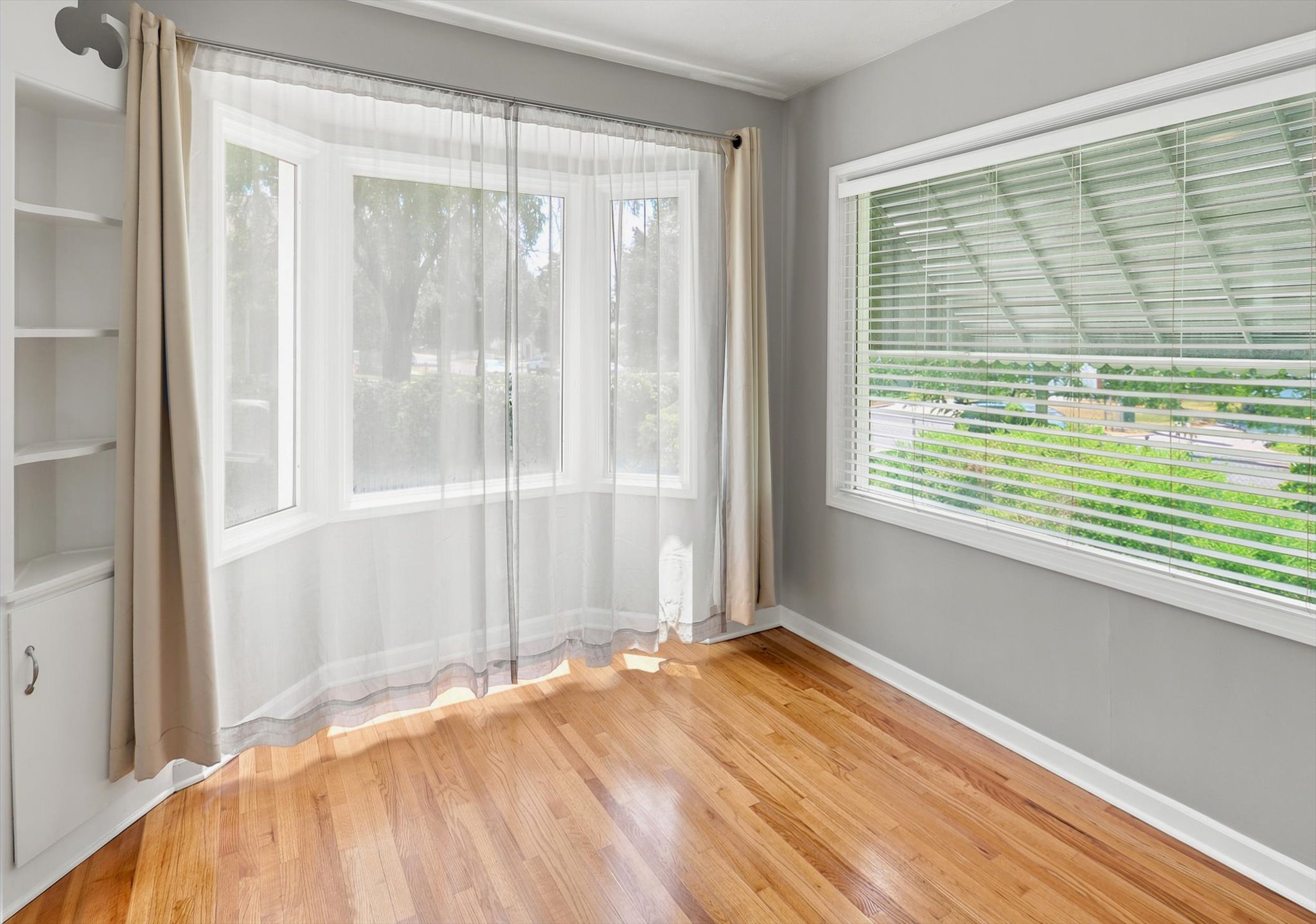 Hardwood flooring throughout this room, featuring a bay window with sheer curtains, a second window with horizontal blinds, built-in shelving, and a built-in cabinet