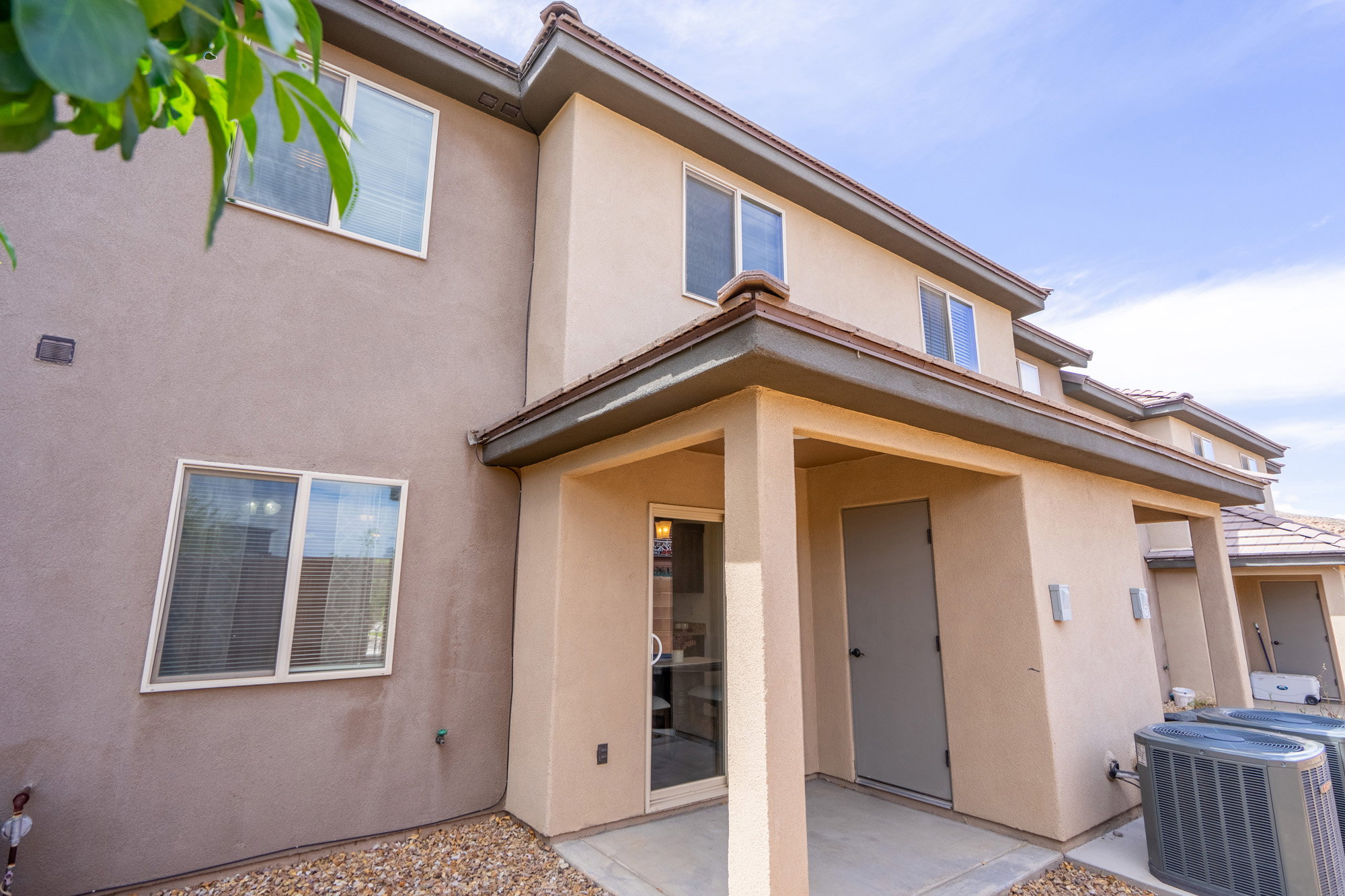 Multi-level stucco exterior with a covered patio area, featuring a sliding glass door, a private storage door, and gravel landscaping