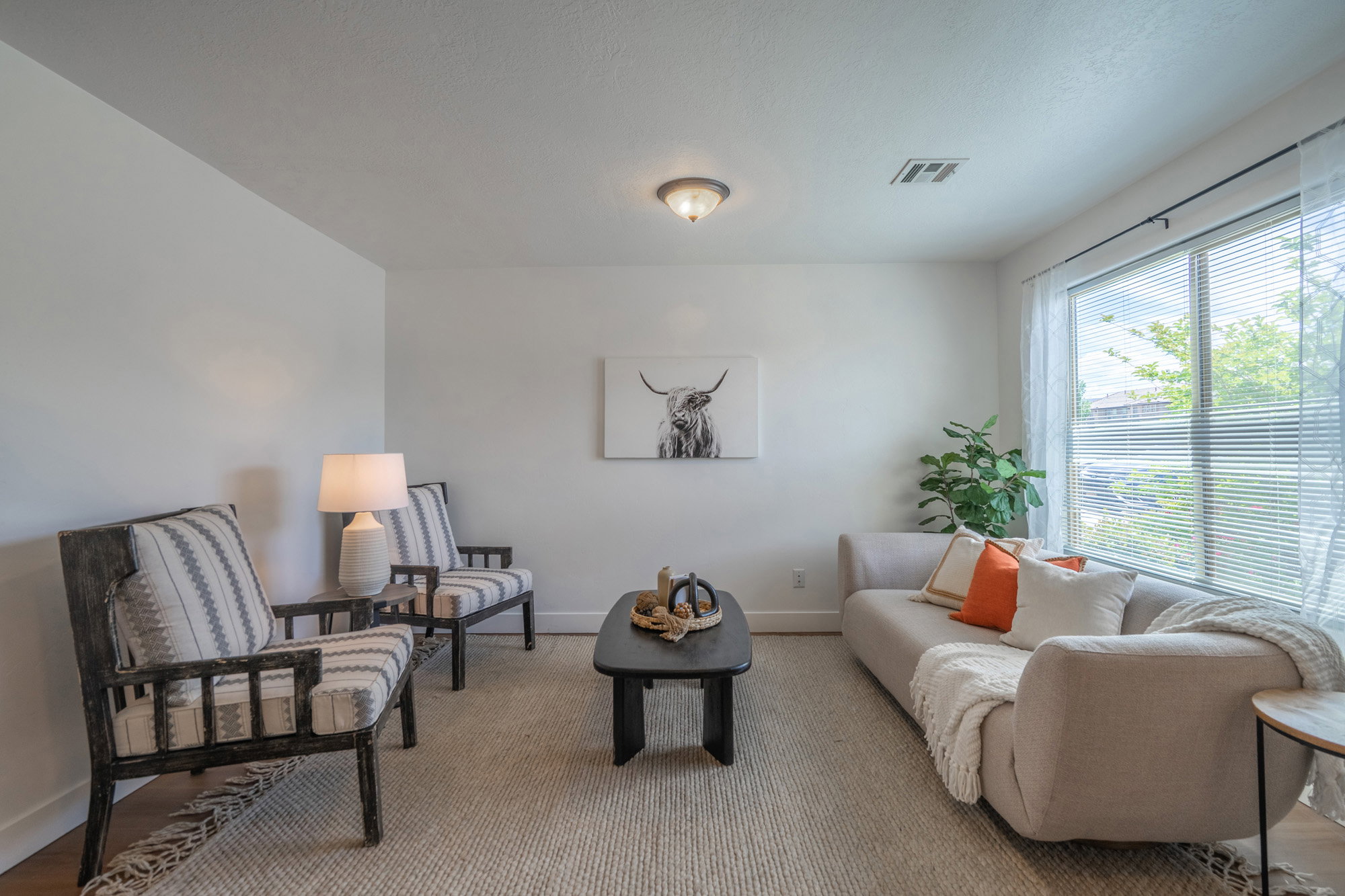 Light-filled living area featuring a broad window with blinds, neutral wall tones, recessed ceiling lighting, and wood-finish flooring partially covered by a textured area rug