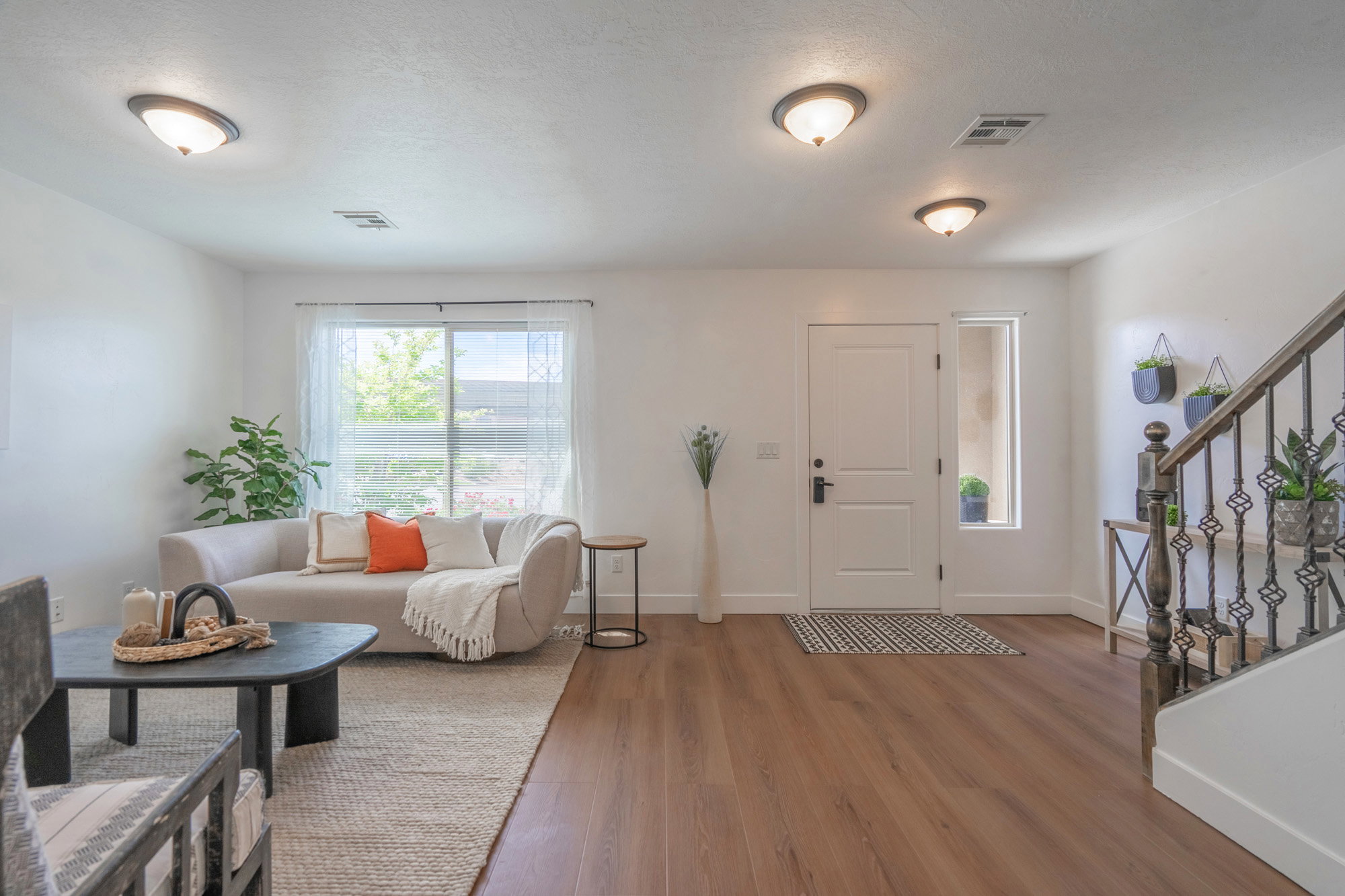 Open-concept living space featuring wood-finish flooring, a white paneled entry door with a sidelight, and a staircase with a dark wood handrail and decorative wrought iron balusters