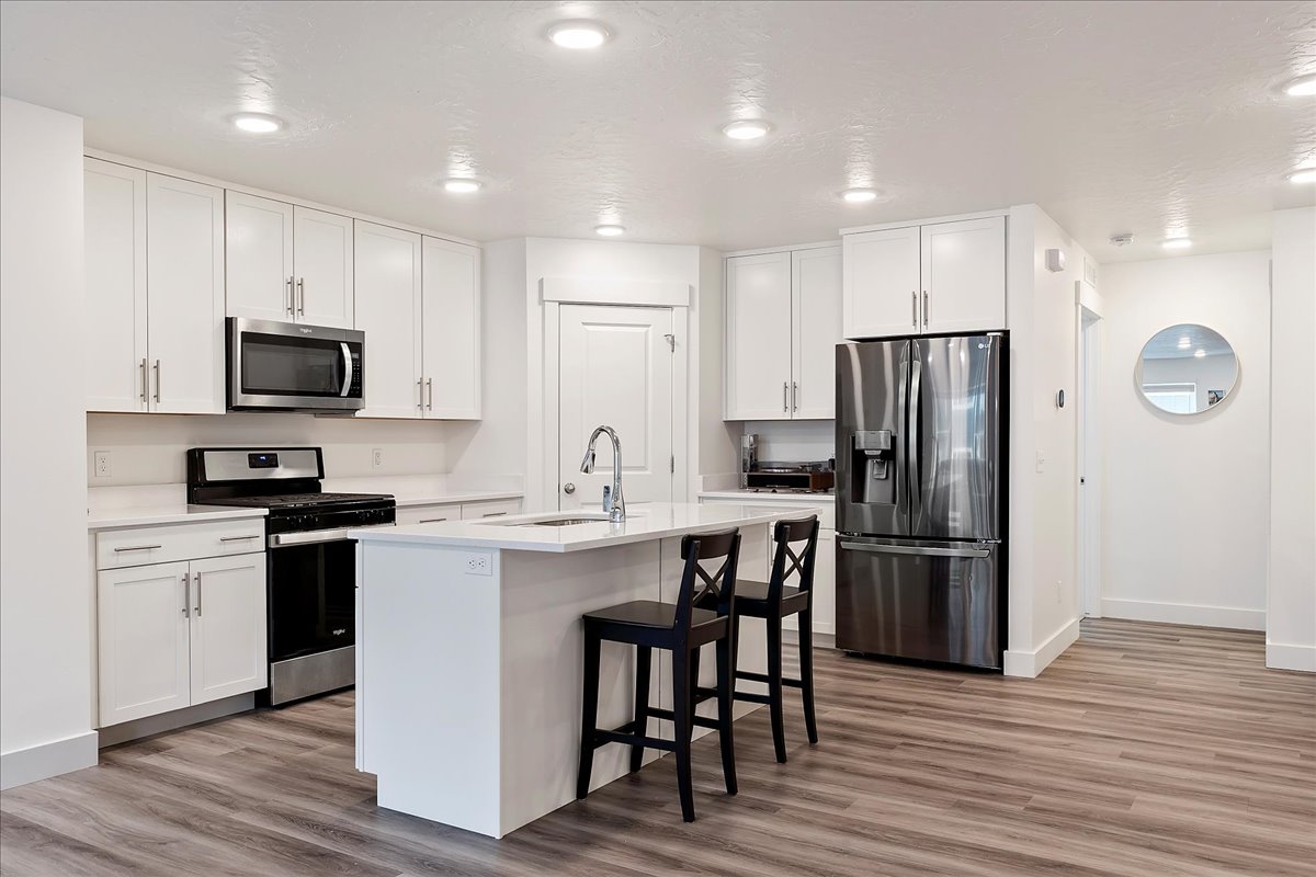 Open-concept kitchen featuring white cabinetry, stainless steel appliances, a center island with sink, quartz countertops, and wood-finish flooring