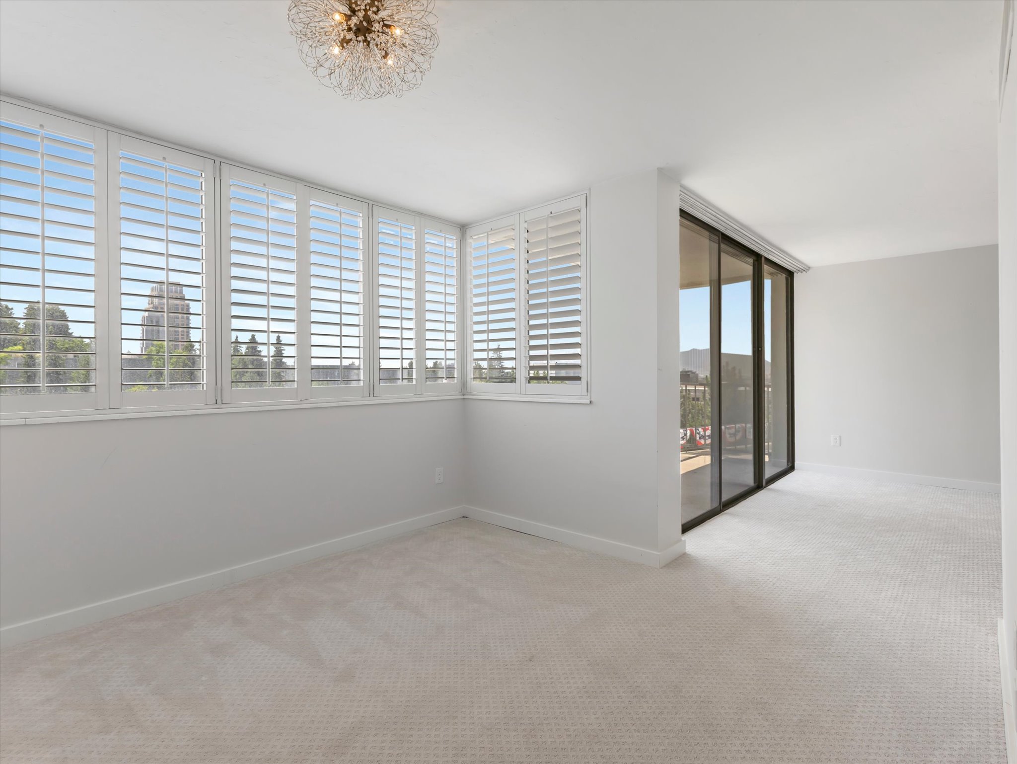 Carpeted interior space featuring a full wall of windows with white plantation shutters, a modern chandelier, and sliding glass doors with dark frames