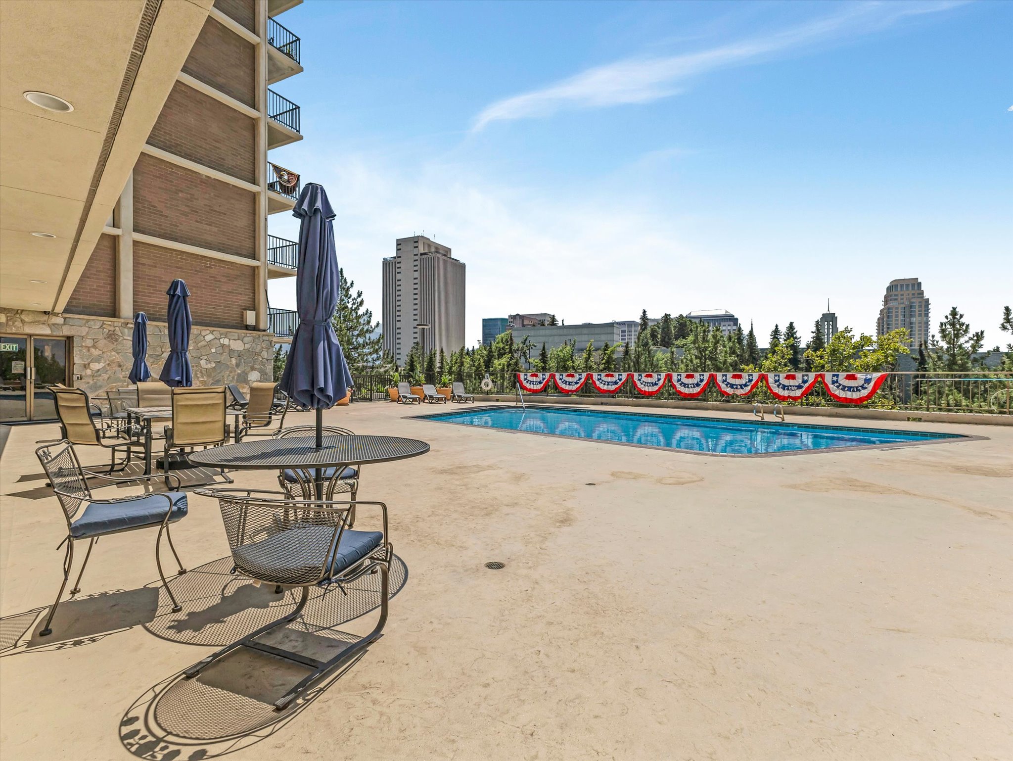 Outdoor swimming pool with expansive concrete decking, multiple seating areas with umbrellas, and a surrounding metal fence