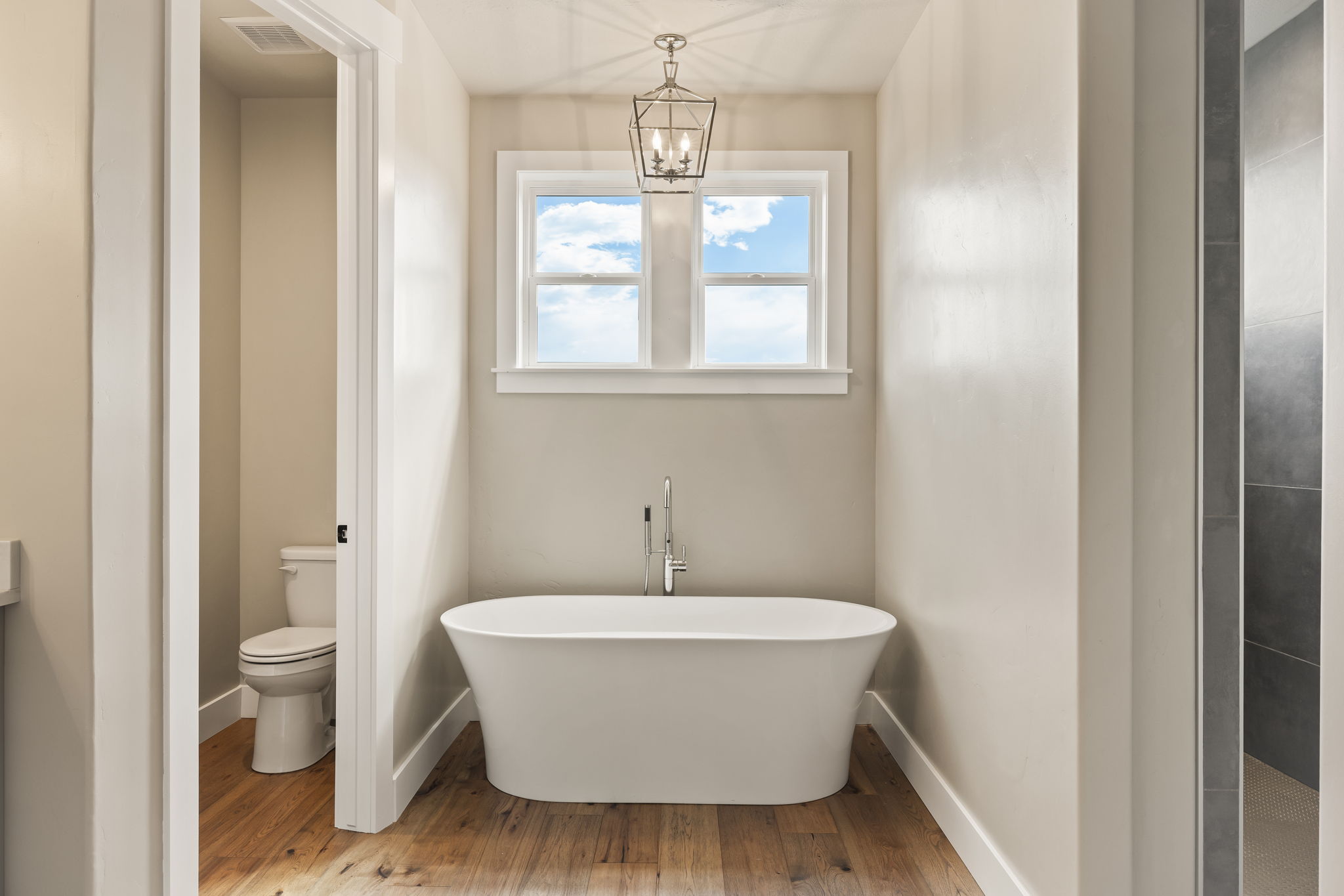 Freestanding soaking tub with a floor-mounted faucet, wood-finish flooring, and a double-pane window
