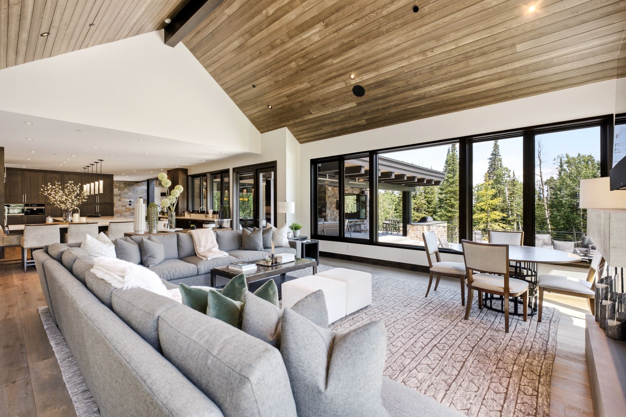 Spacious open-concept living area featuring a vaulted wood-finish ceiling, large black-framed picture windows showcasing exterior greenery, and wood-finish flooring