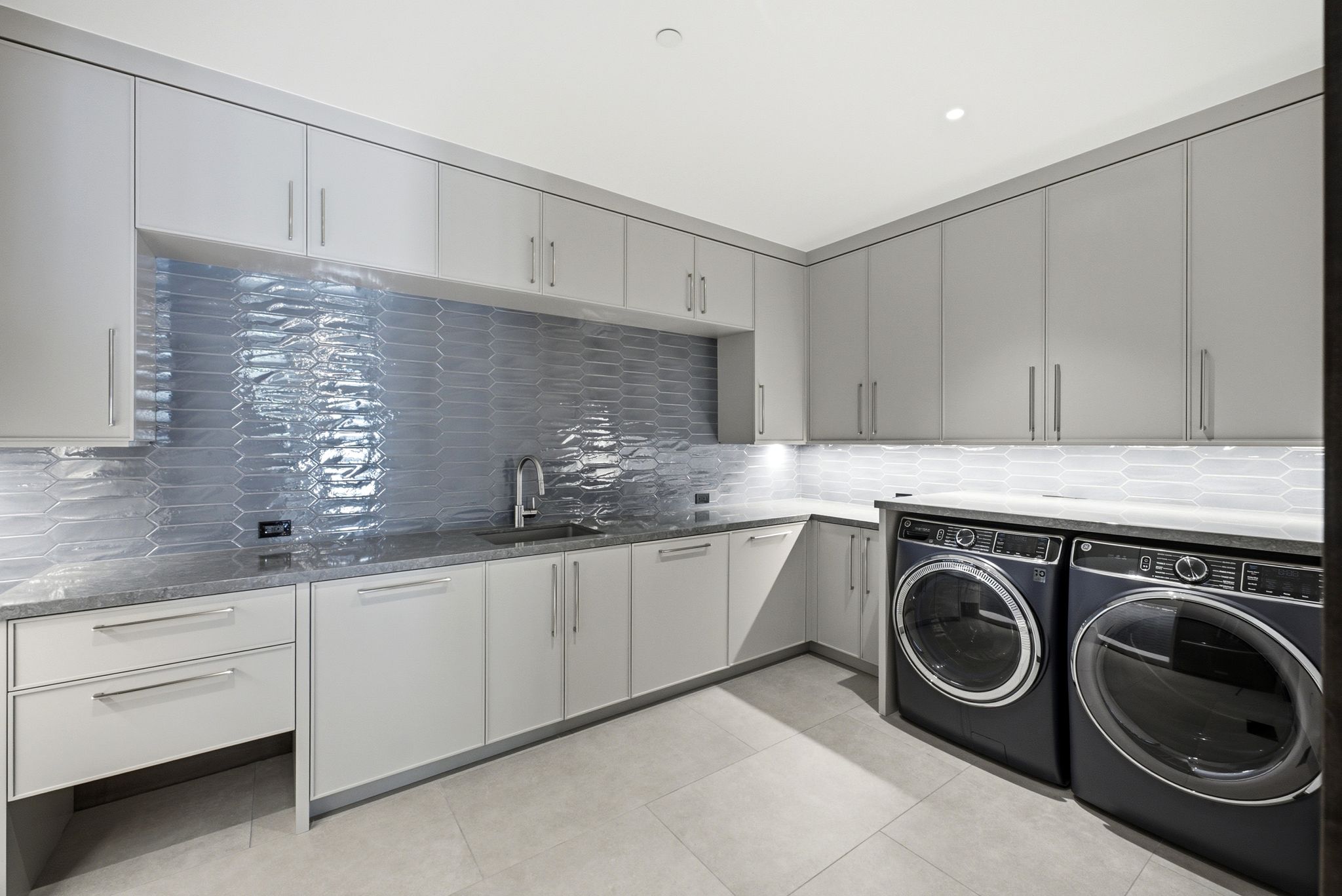 Modern laundry area featuring an under-mount sink, extensive cabinetry, light-toned tile flooring, and under-cabinet lighting