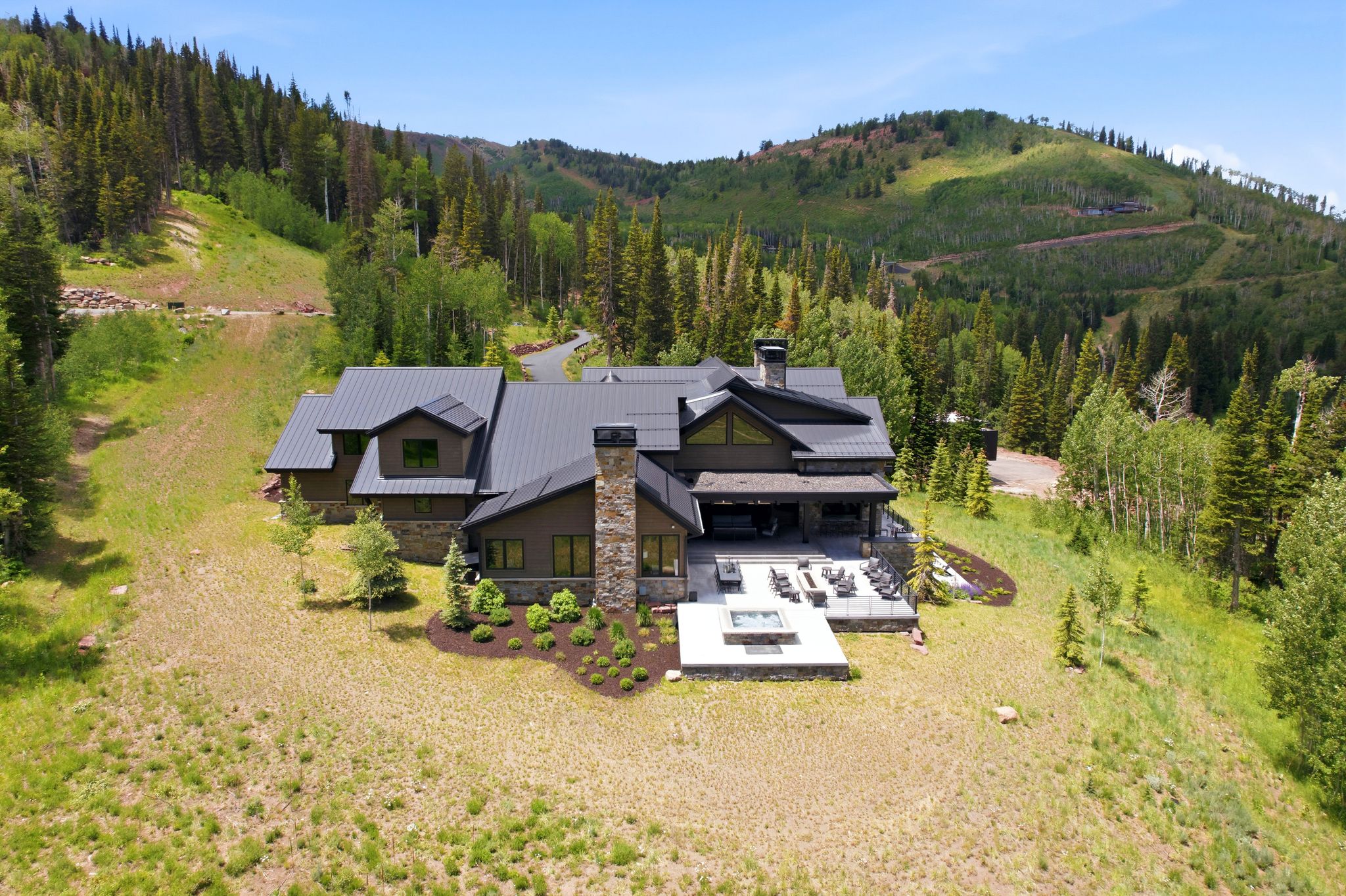 Contemporary mountain residence featuring a dark metal roof, stone chimney, and expansive outdoor patio with fire pit