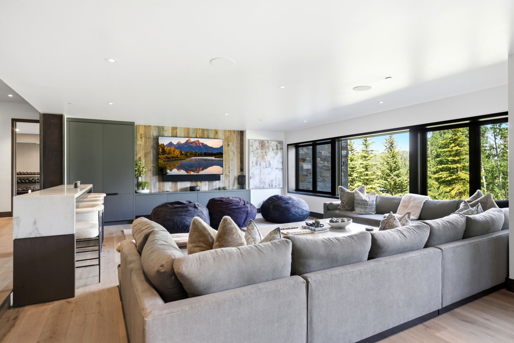 Spacious media room featuring wood-finish flooring, a built-in wet bar with a stone-finish countertop, a reclaimed wood-paneled accent wall, and expansive windows with dark frames