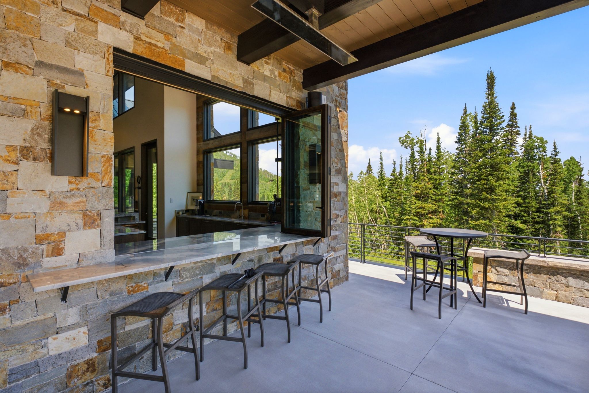 Outdoor entertaining space featuring natural stone veneer, a pass-through bar with a light-toned countertop, a folding window system, and a concrete patio with a cable railing system overlooking evergreen trees