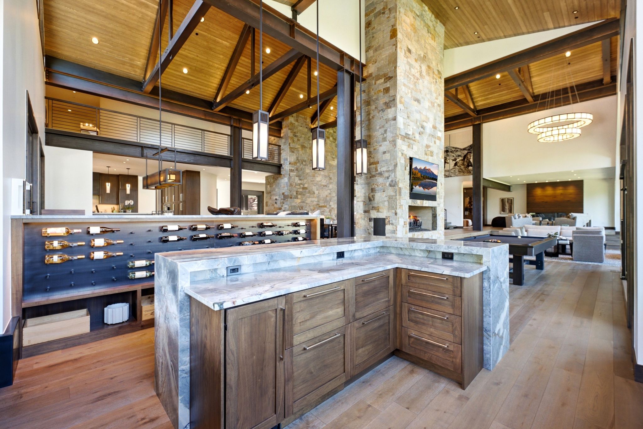 Vaulted wood-paneled ceilings with exposed beams, stone-clad fireplace with built-in media, integrated wine storage, light wood-finish flooring, and a built-in bar with stone countertops and wood cabinetry
