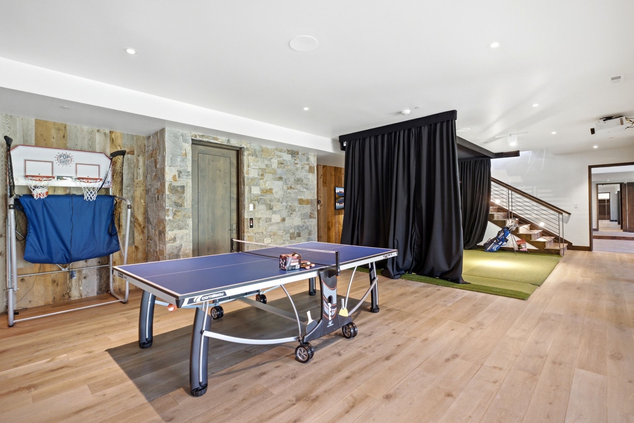 Spacious recreation area featuring wood-finish flooring, stone accent wall, recessed lighting, and a golf simulator area with black curtain enclosure