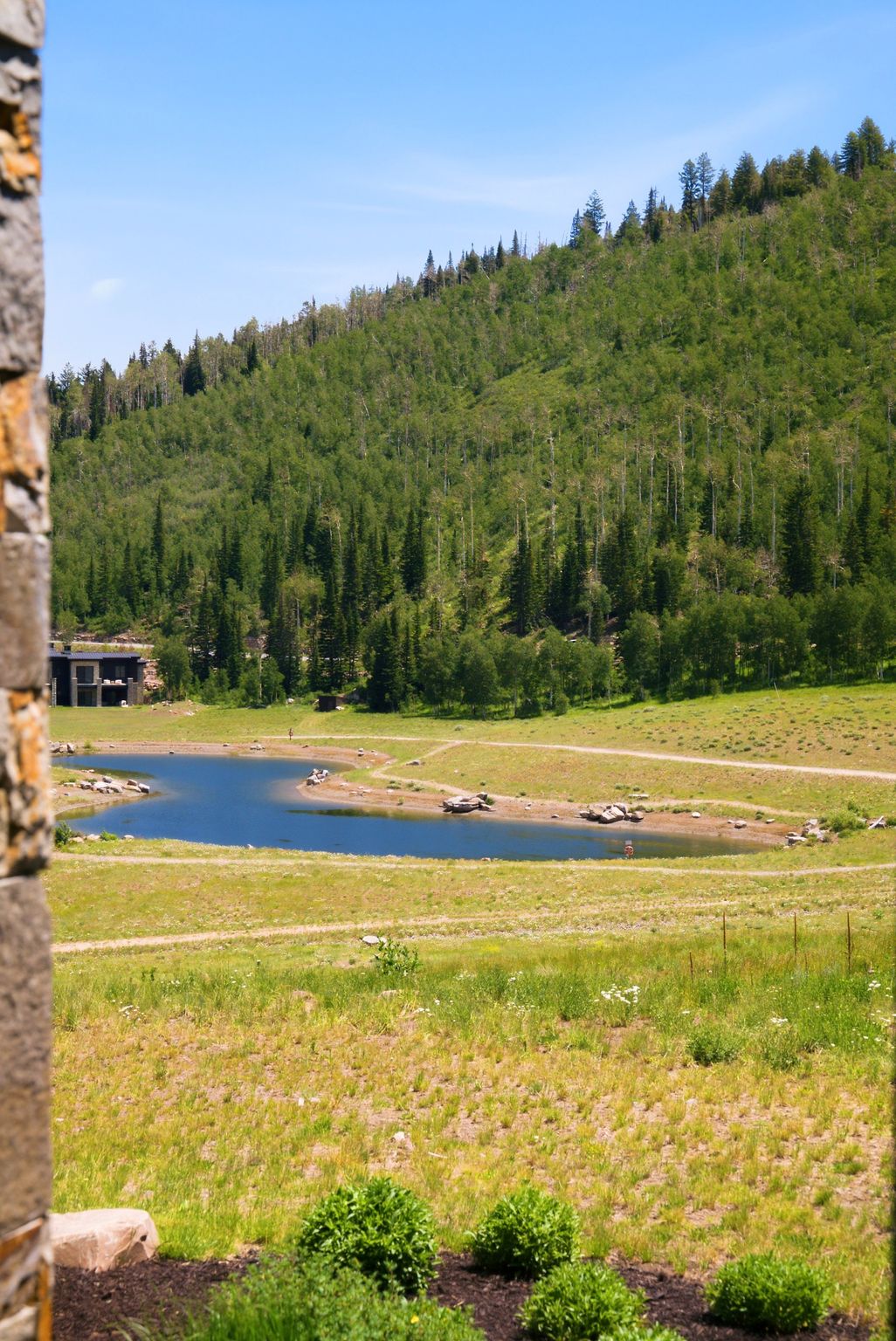 Expansive natural landscape featuring a prominent blue pond, surrounded by grassy terrain and a backdrop of verdant forested hills