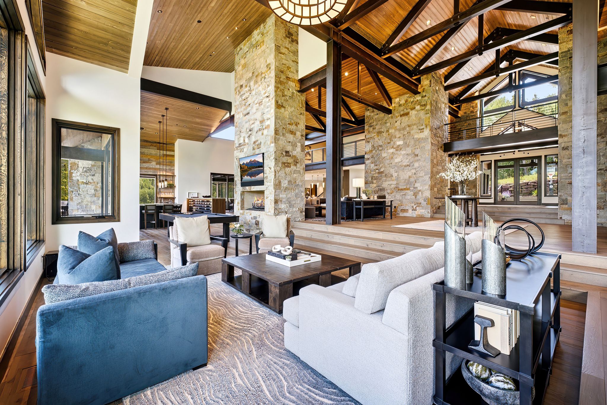 Vaulted wood-paneled ceilings with exposed beams, stone accent walls, expansive windows, and tiered wood-finish flooring define this grand living space