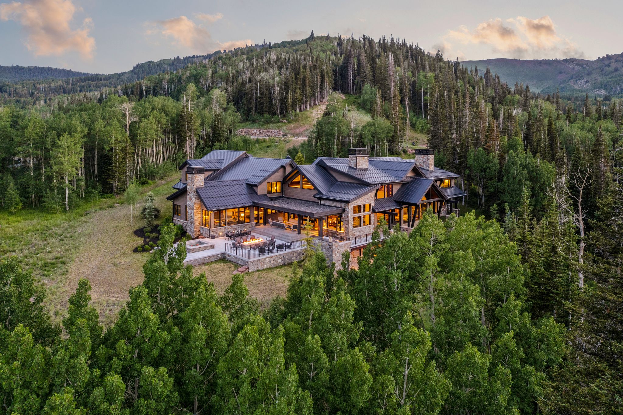 Stone and timber frame mountain residence featuring a multi-level exterior, dark metal roofing, extensive outdoor patio space, and a stone fire pit