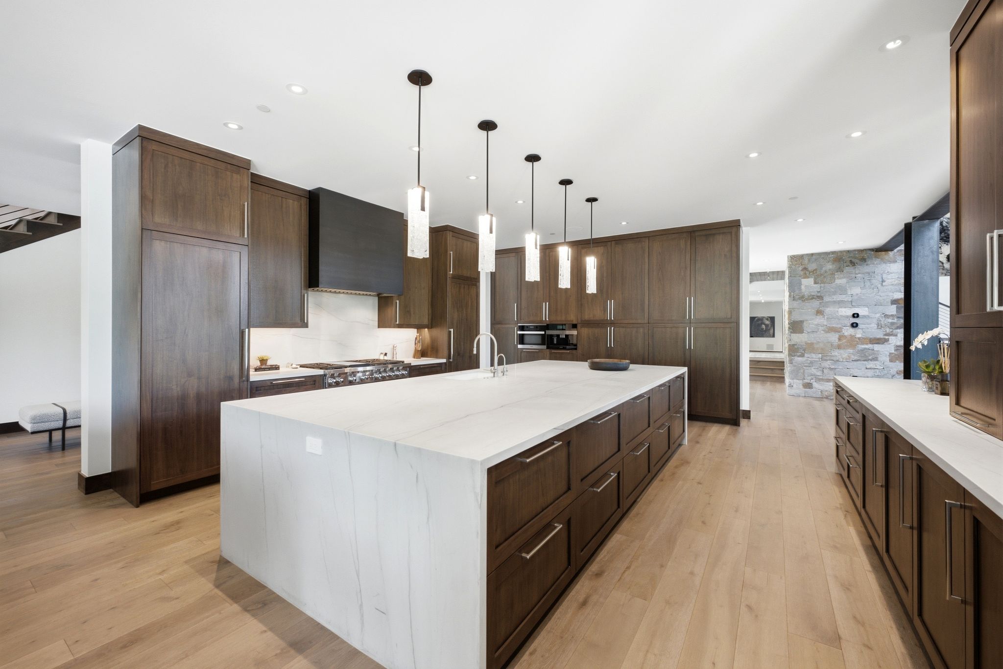 Contemporary kitchen featuring a large waterfall island with a white stone countertop, extensive dark wood cabinetry, a professional-grade range with a built-in hood, and light wood-finish flooring