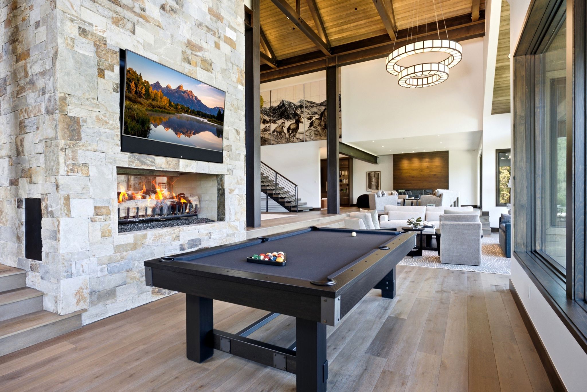 Spacious living area featuring a stone-clad fireplace with built-in television, wood-finish flooring, vaulted ceilings with exposed beams, large windows, and contemporary light fixtures