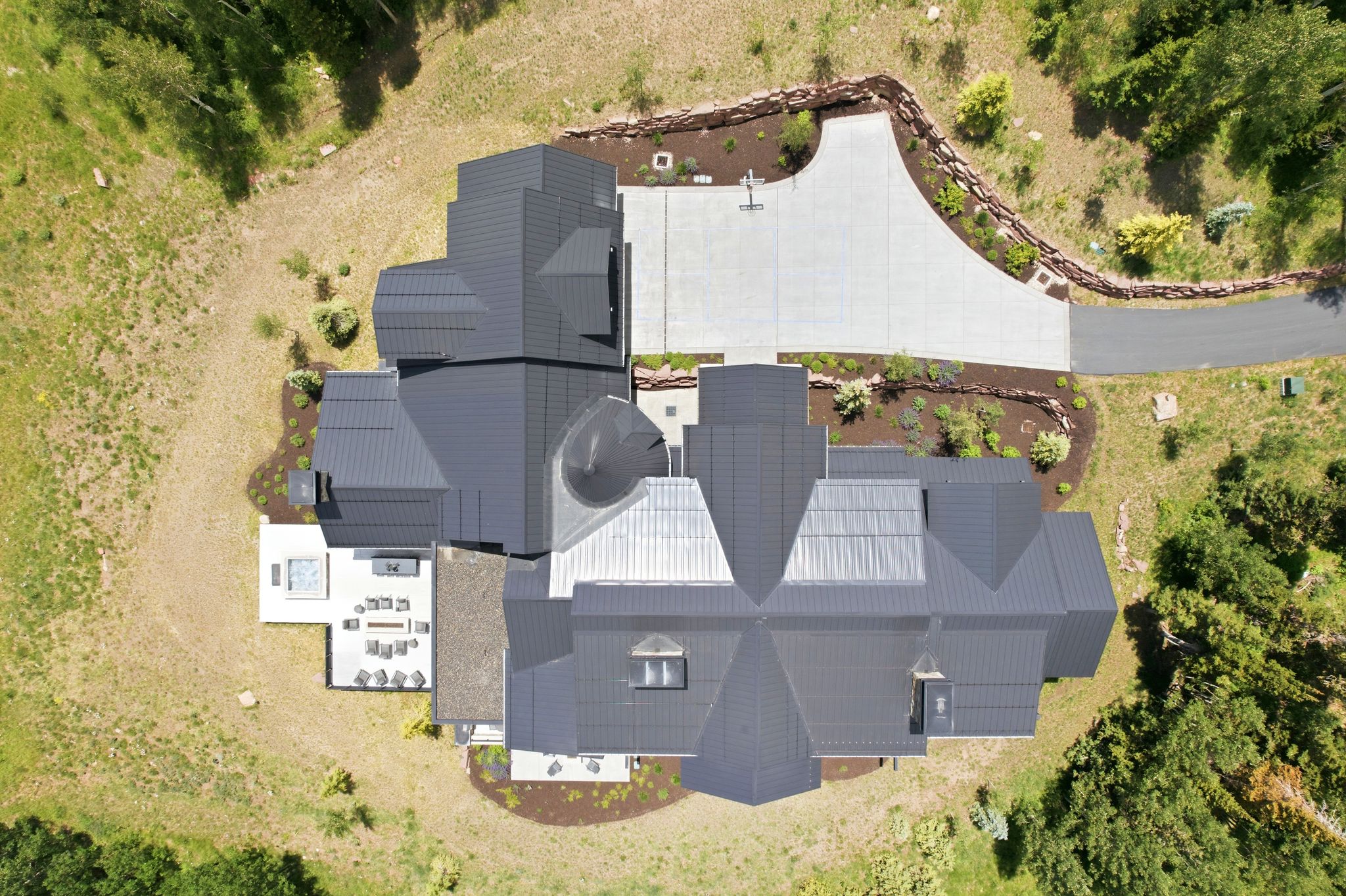Expansive property featuring a multi-level roofline, a large paved driveway, an outdoor patio with a visible built-in hot tub, and surrounding natural landscaping