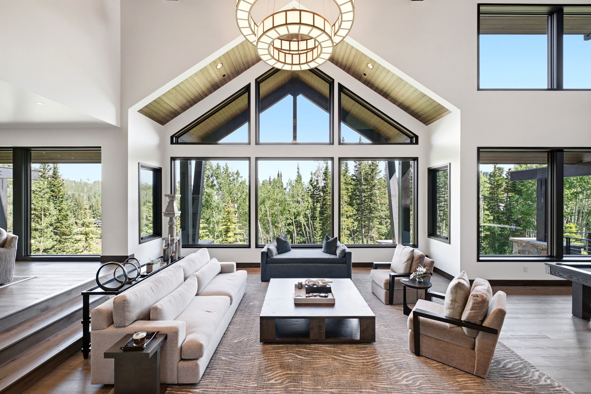 Spacious living area featuring a vaulted ceiling with wood-paneled accents, a large circular chandelier, and extensive fenestration with black frames