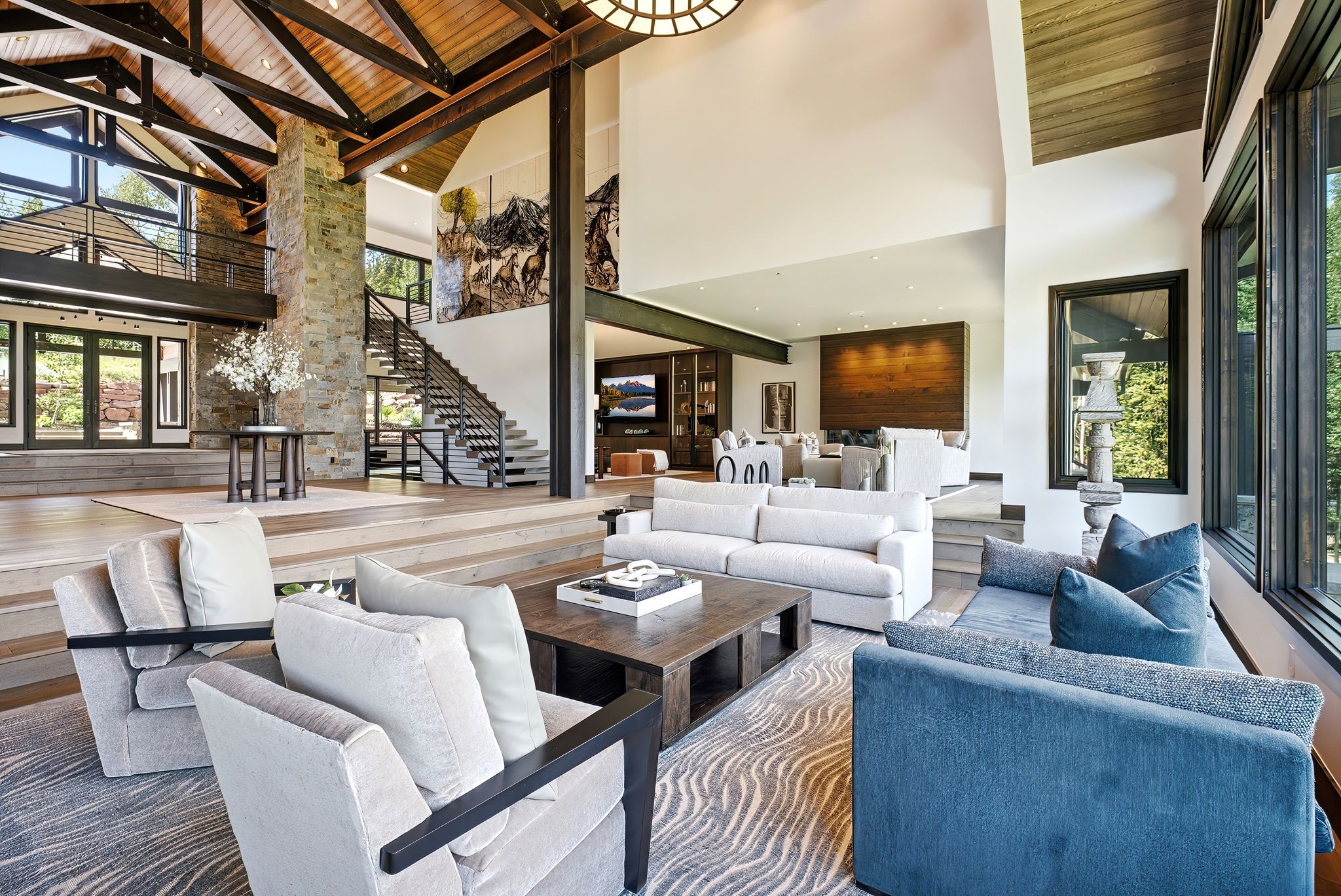 Grand living area featuring soaring vaulted ceilings with exposed wood beams, a stone-clad fireplace column, multi-level wood-finish flooring, a contemporary open-riser staircase with metal railings, and expansive floor-to-ceiling windows