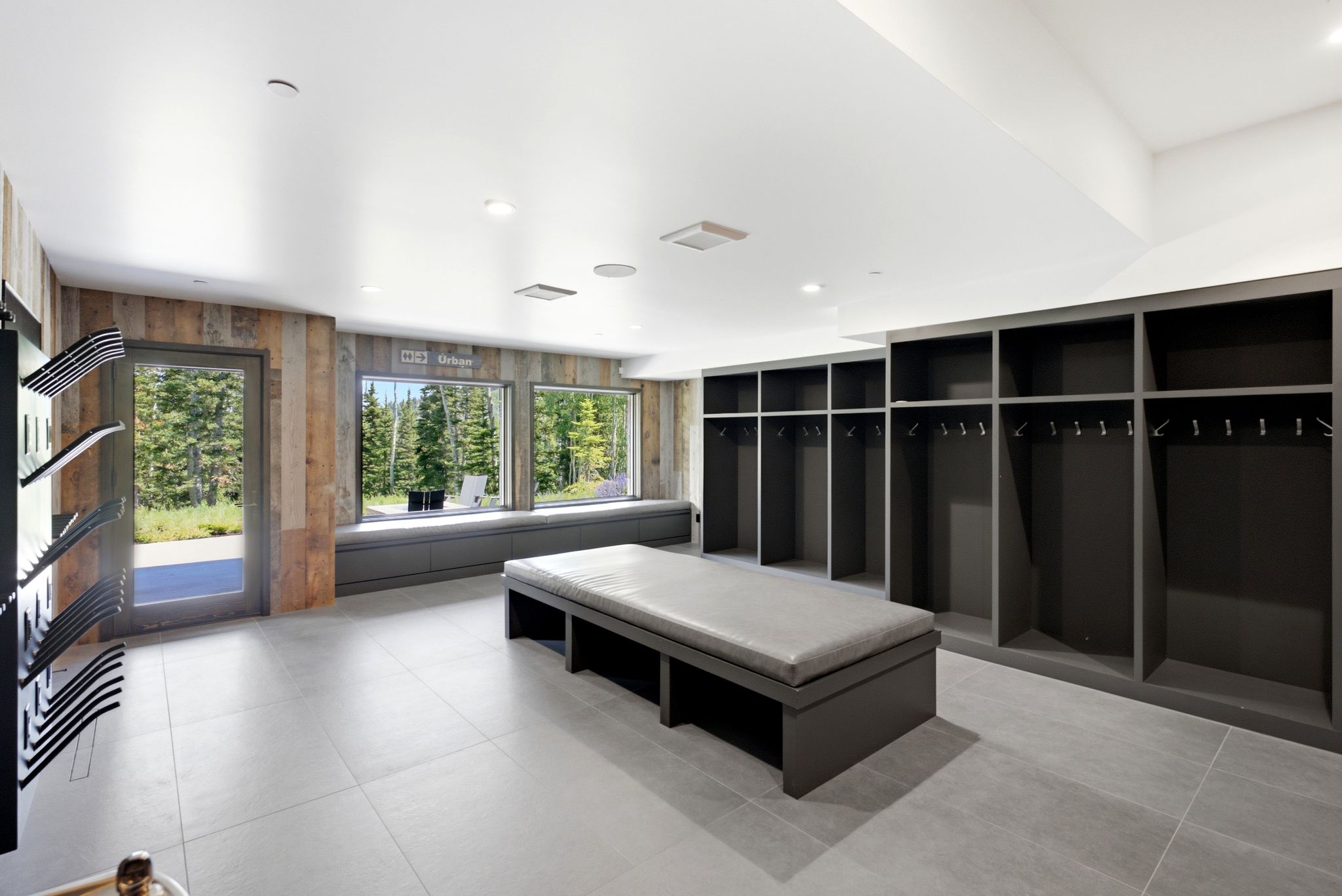 Spacious room featuring light gray tile flooring, extensive built-in open lockers with hanging rods, a central upholstered bench, and a wall of windows showcasing natural scenery