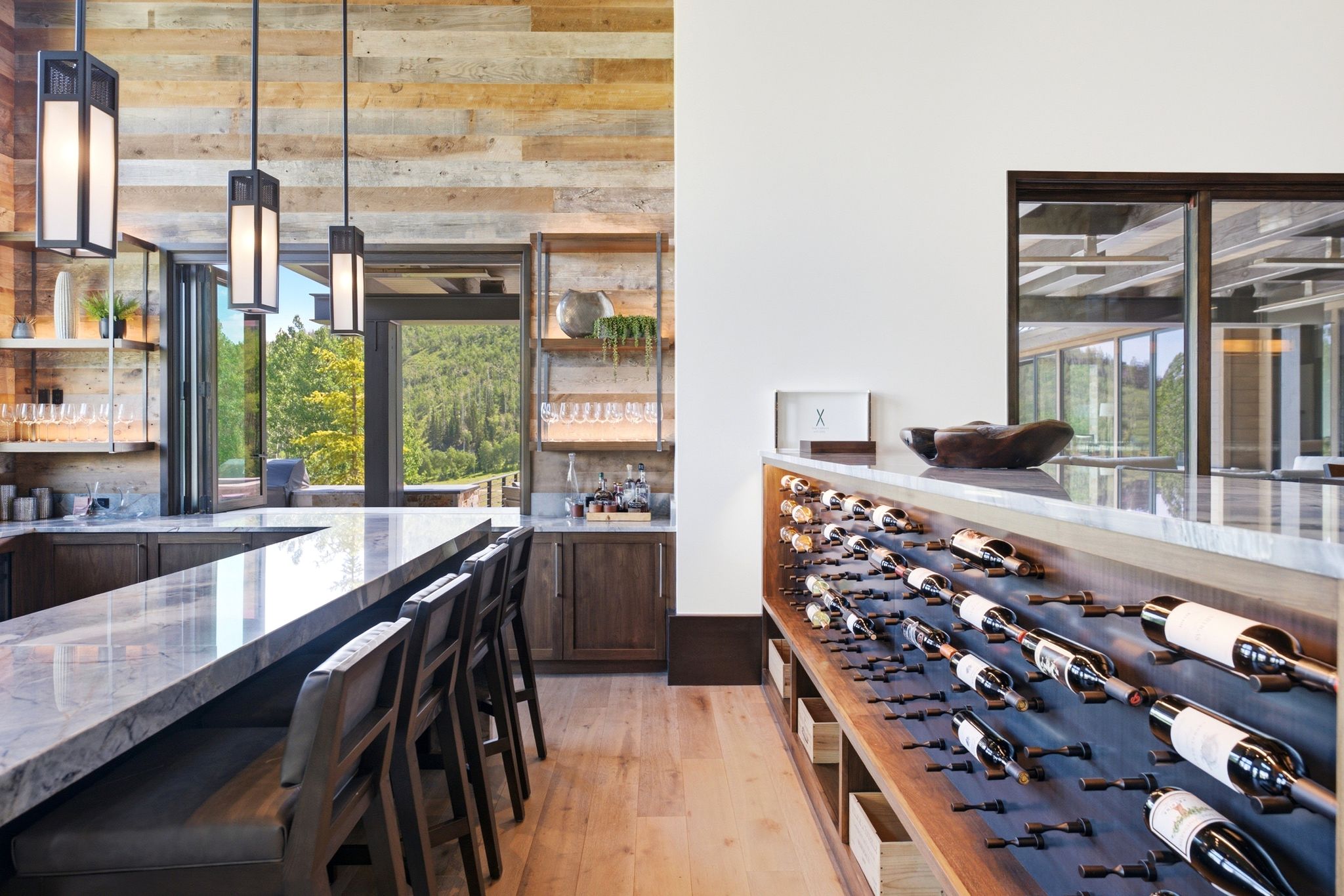 Entertainment area featuring a wood-finish accent wall, a stone-topped bar with seating, a built-in wine storage display, and bi-fold glass doors opening to a scenic mountain backdrop