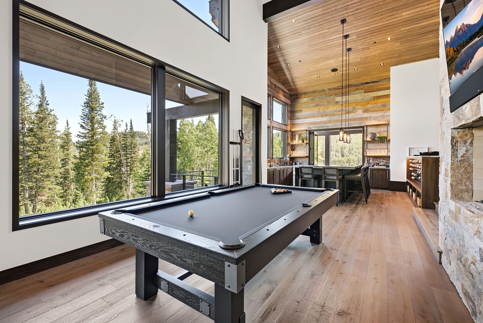 Spacious entertainment area featuring expansive windows, wood-finish flooring, a vaulted wood-paneled ceiling, a built-in wet bar with seating, and a stone-clad fireplace surround