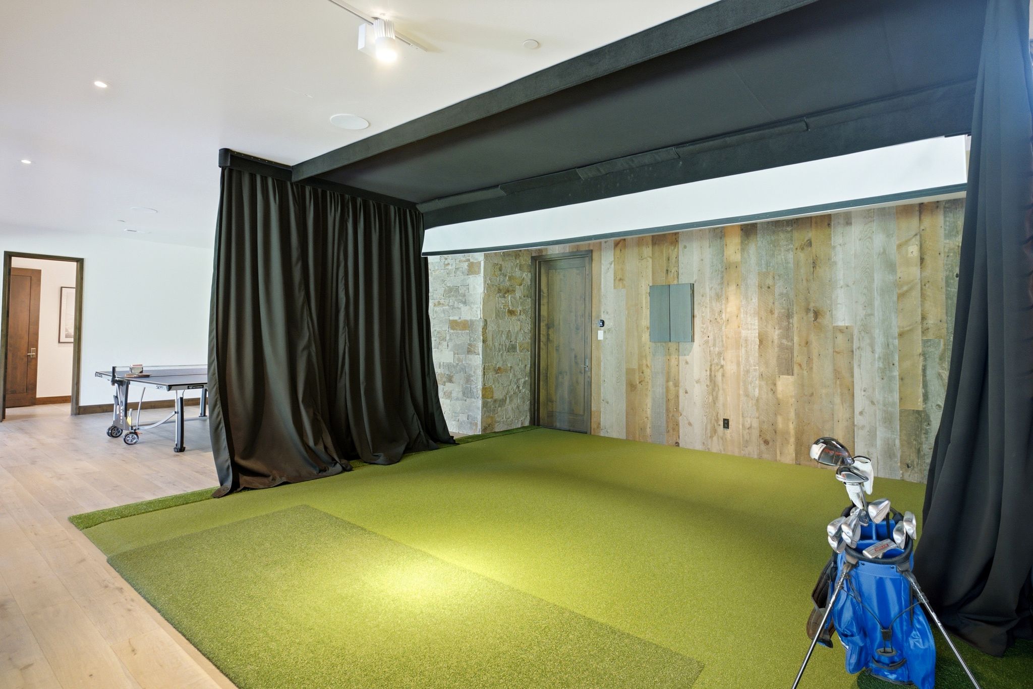 Indoor golf simulation area featuring turf flooring, full-height privacy curtains, and a reclaimed wood accent wall