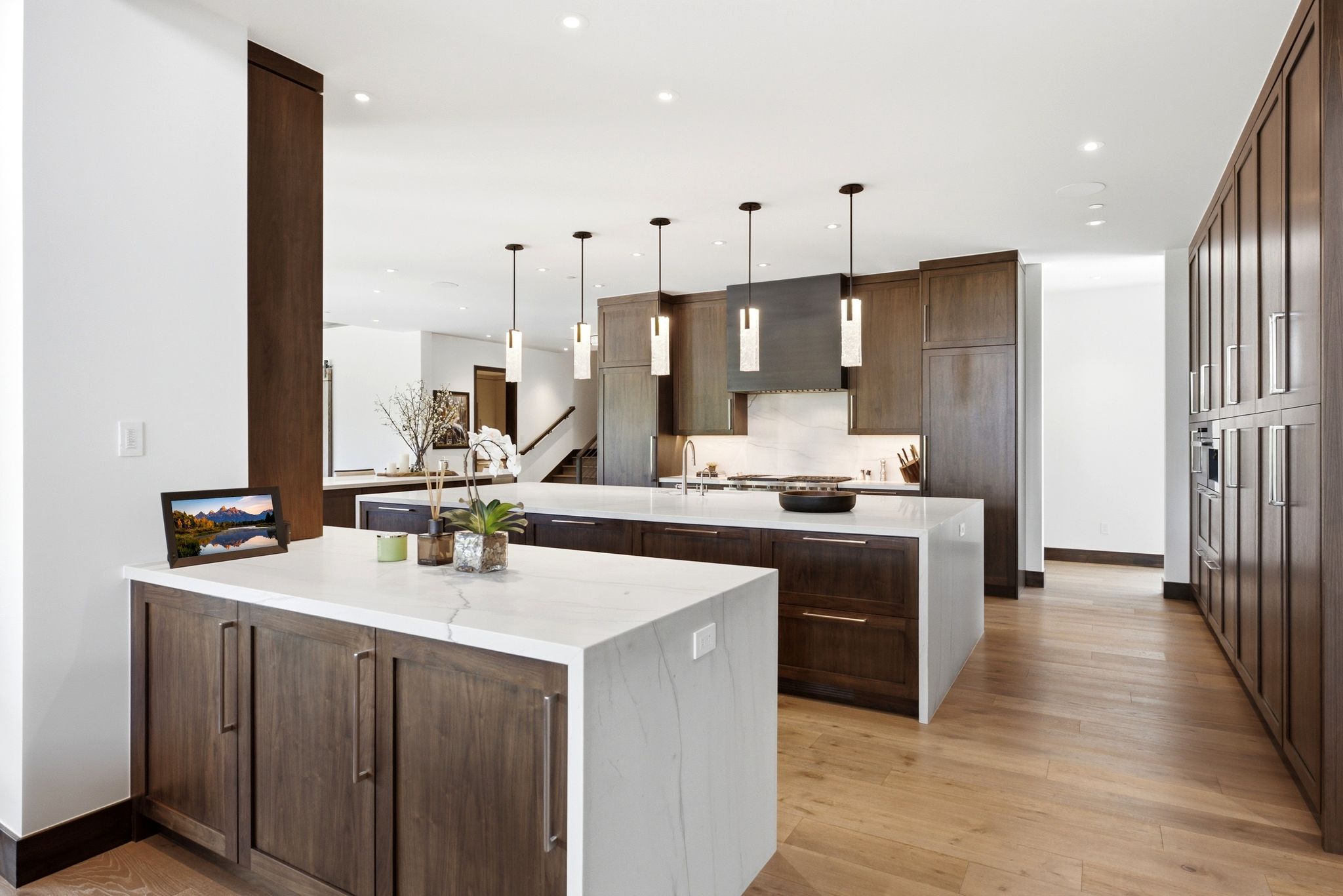 Spacious kitchen featuring two large islands with waterfall countertops, extensive dark wood-finish cabinetry, wood-finish flooring, recessed lighting, and modern pendant fixtures