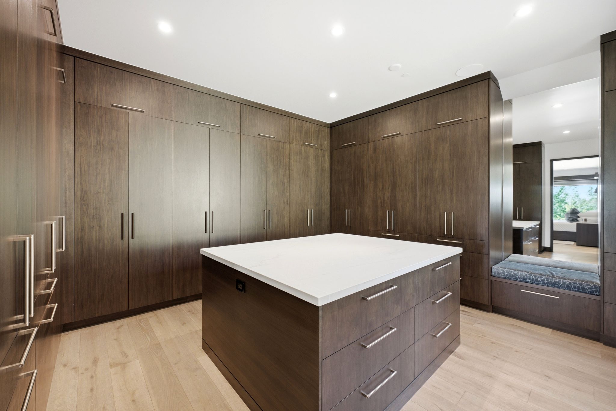 Spacious dressing area featuring extensive dark wood-finish cabinetry with brushed nickel hardware, a central island with a white countertop and drawer storage, light wood-finish flooring, and recessed ceiling lighting