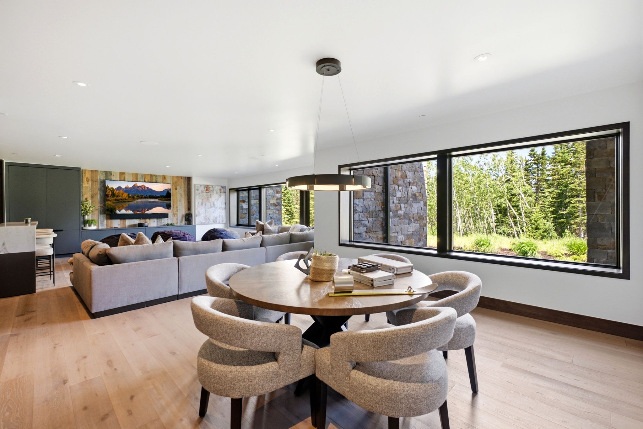 Spacious open-concept living area featuring light wood-finish flooring, a contemporary circular chandelier, and expansive windows showcasing exterior foliage