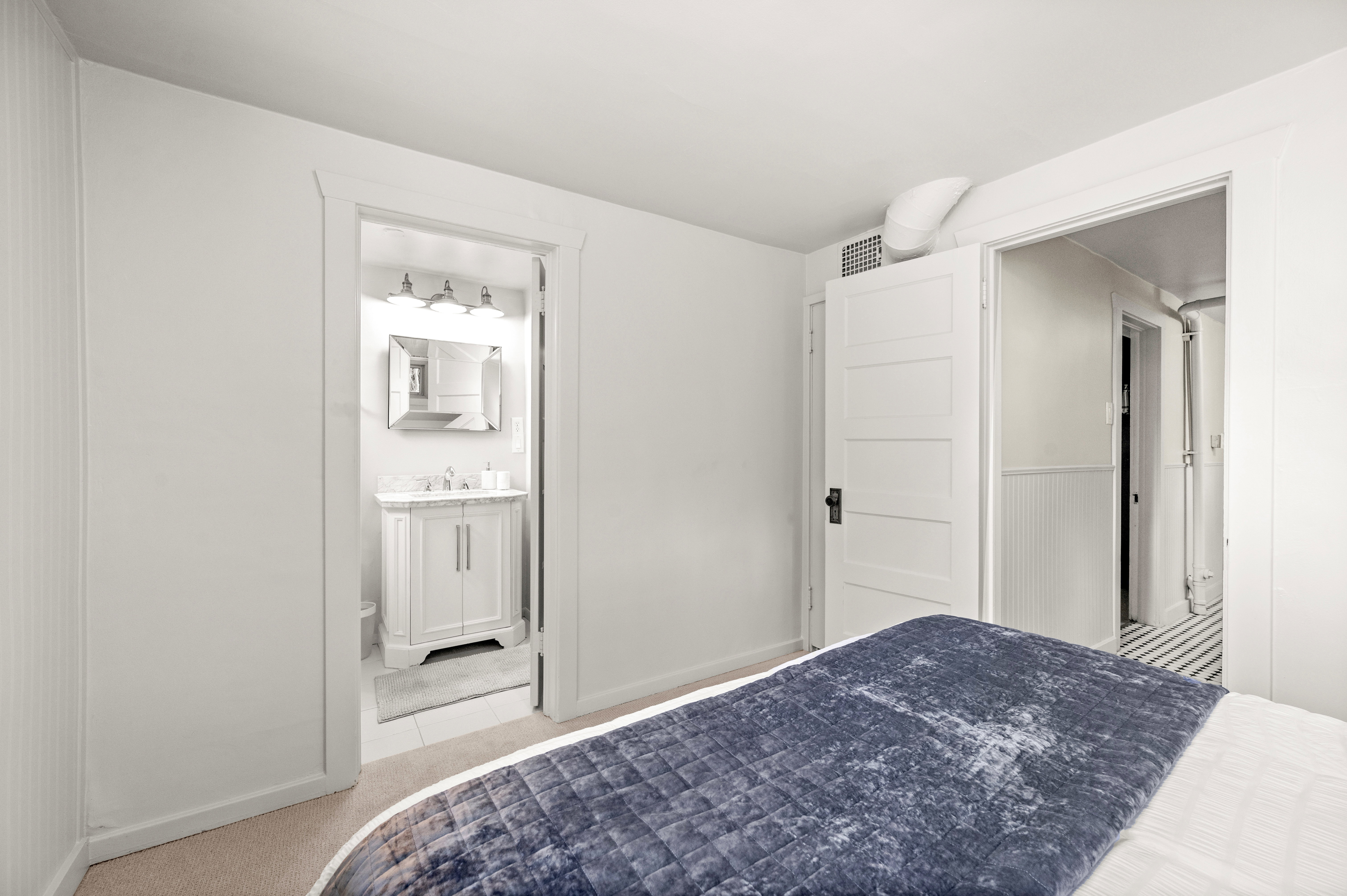 White interior walls featuring a traditional paneled door, light-colored carpeting, and a visible bathroom entry with a white vanity and wall-mounted mirror