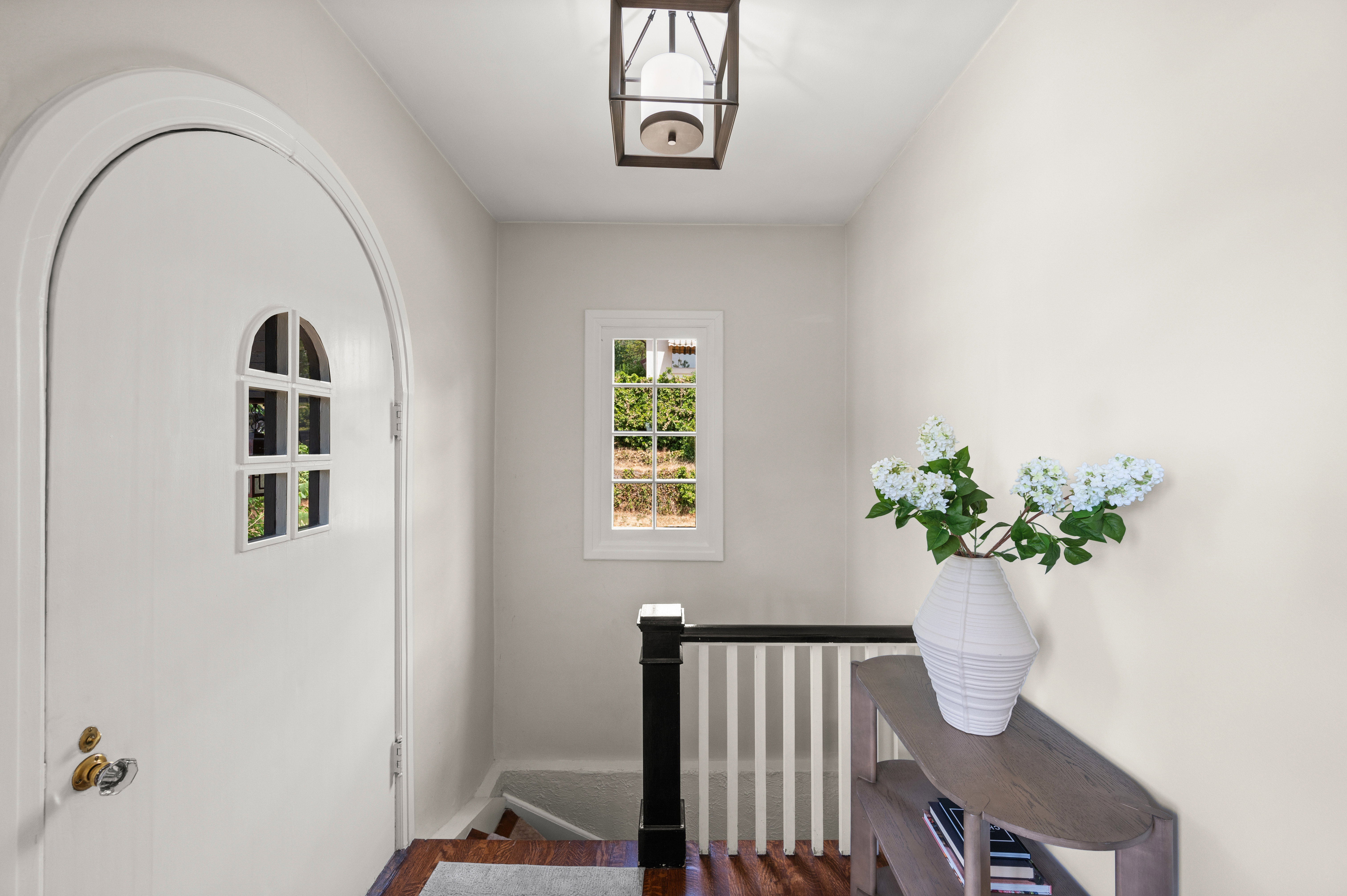 Arched doorway with an integrated window, featuring wood-finish flooring, a contemporary ceiling lantern, and a classic black and white staircase railing