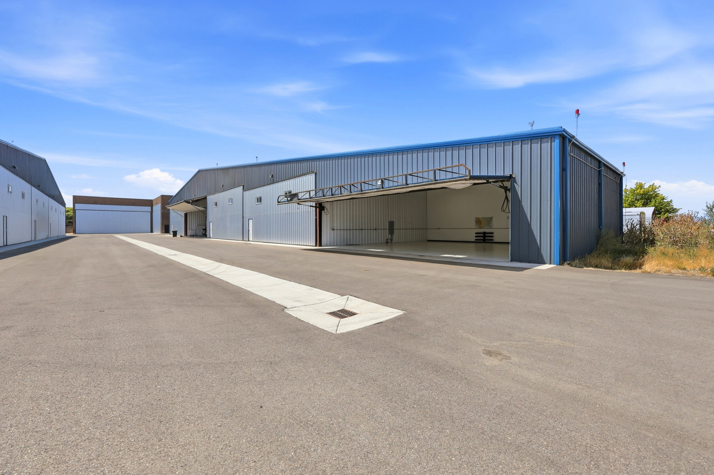 Industrial metal building with blue trim, featuring a large open bay door, paved lot, and additional structures in the background