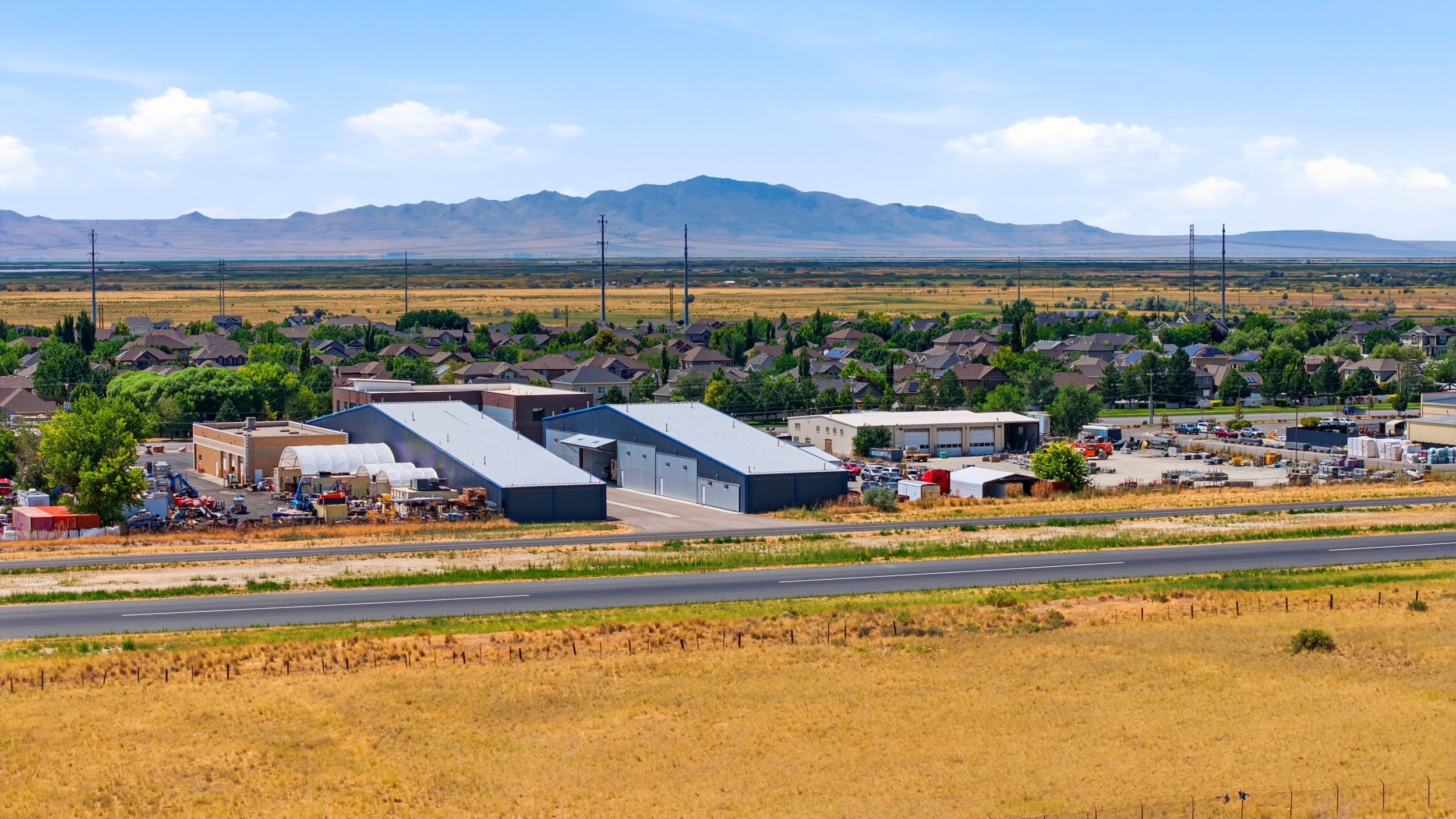 Expansive commercial property featuring multiple metal-clad buildings with roll-up doors, a paved service road, and open land
