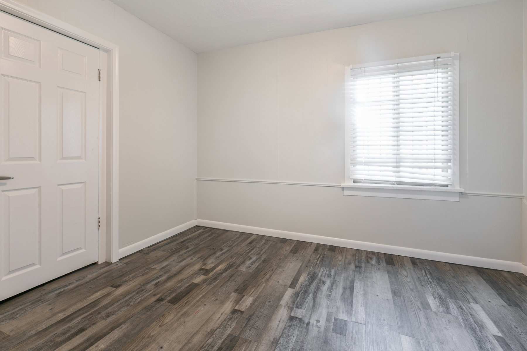 Spacious room featuring wood-finish flooring, light neutral wall tones, a window with blinds, white trim, and a paneled door