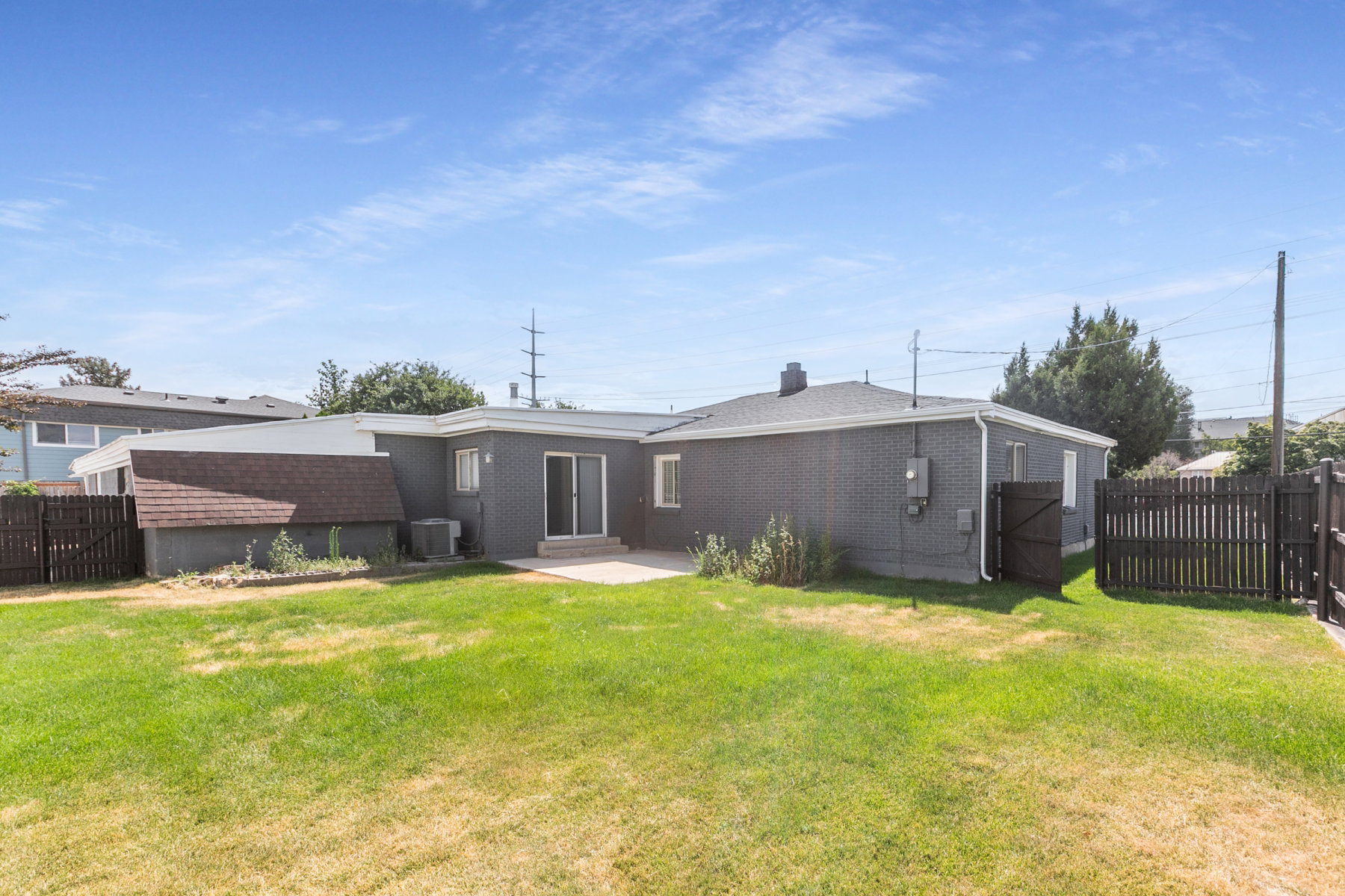 Spacious backyard featuring a large green lawn, a concrete patio, dark gray painted brick siding, a sliding glass door, and dark wood fencing