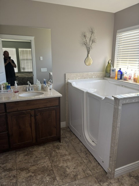 Walk-in tub with textured surround, single vanity with wood cabinetry, light-toned countertop, and patterned floor tile