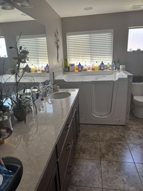 Spacious bathroom featuring a walk-in tub, dual vanity with a light-toned countertop, dark wood-finish cabinetry, and large format floor tiling