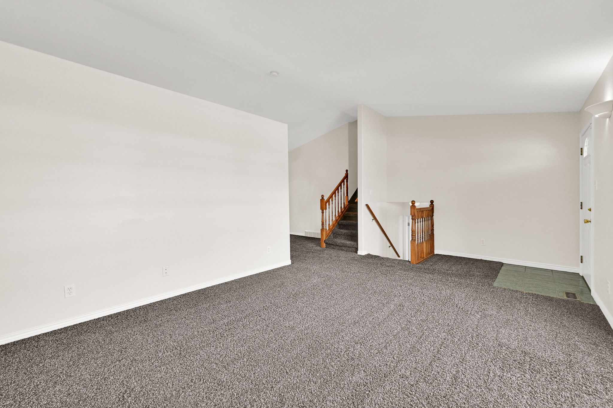 Main living area featuring neutral-toned walls, grey carpet flooring, and a wood staircase with balusters