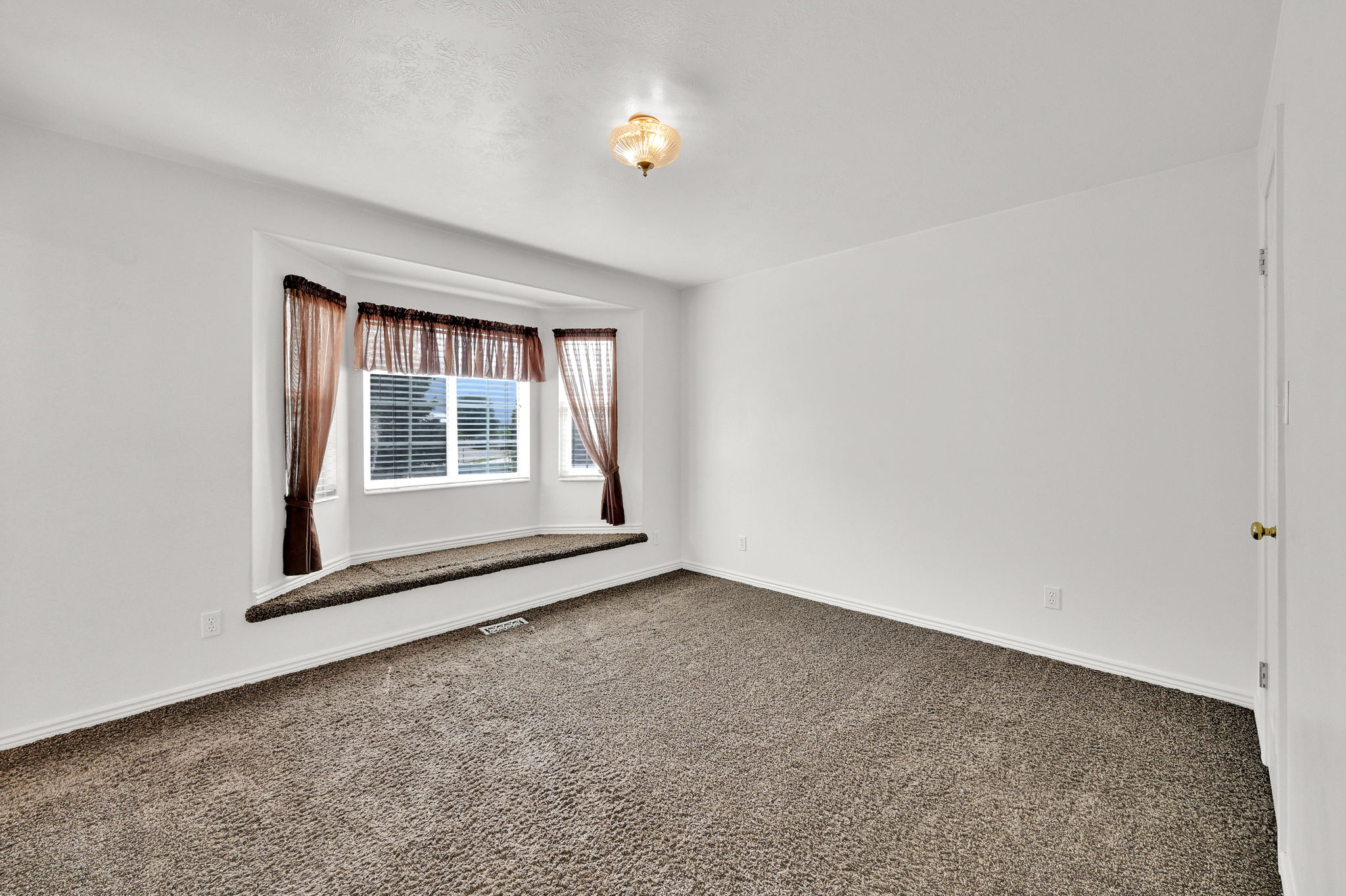 Room with textured carpet flooring and a bay window featuring white blinds and sheer brown curtains