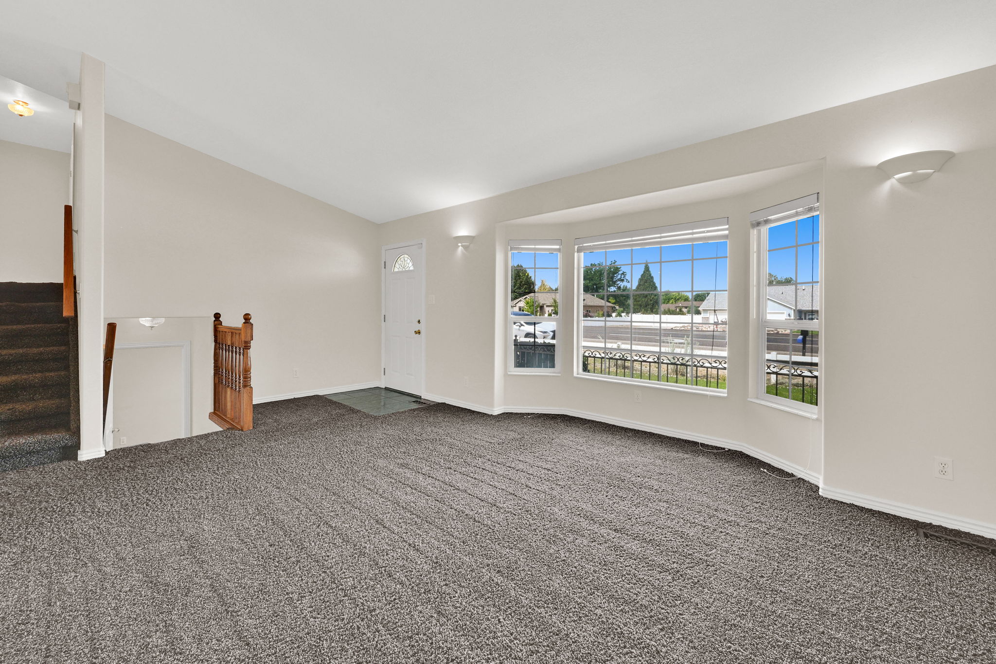 Living area featuring a bay window, wall-mounted sconce lighting, neutral wall paint, and gray textured carpeting