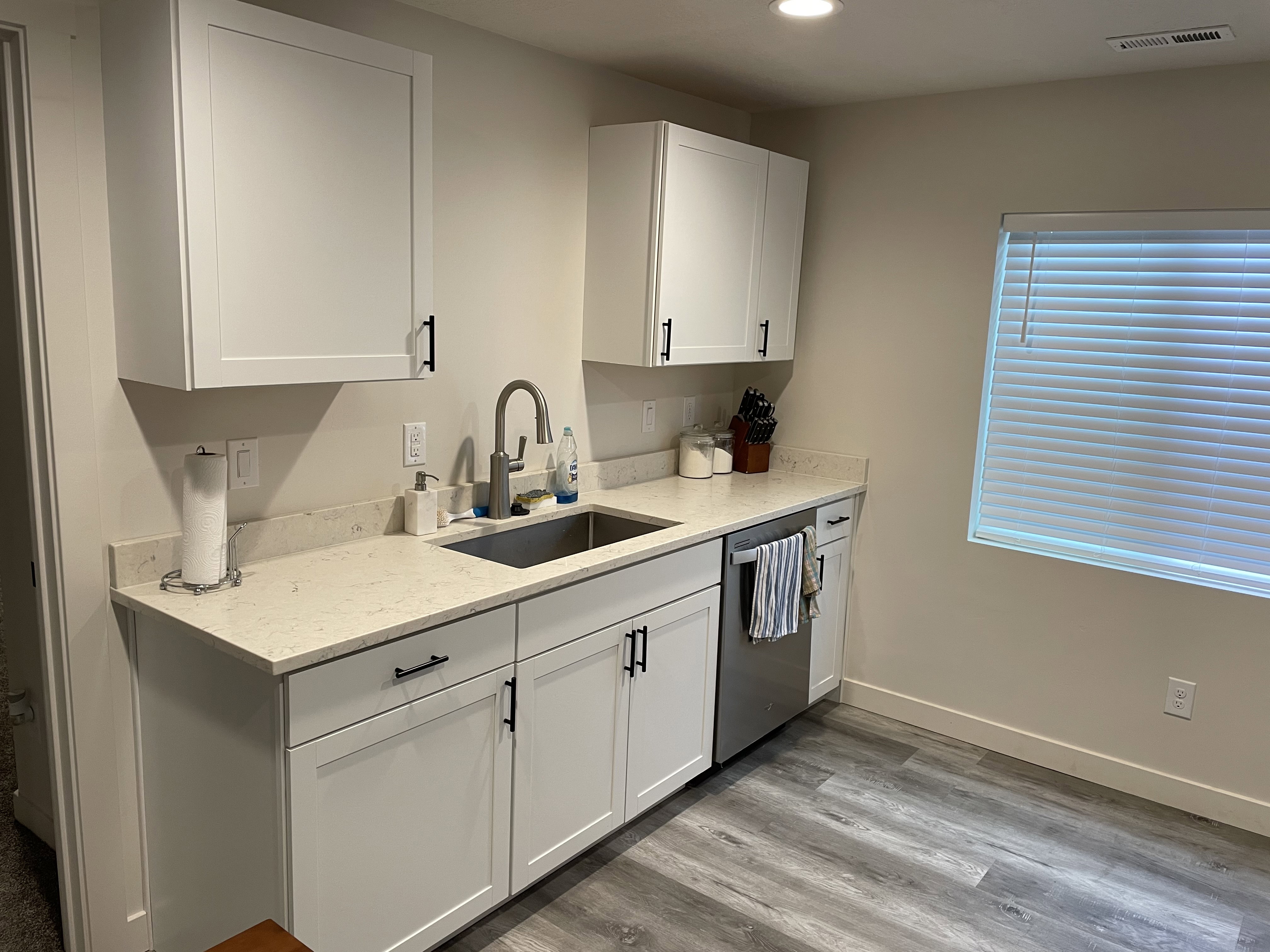 Kitchenette featuring white shaker-style cabinetry, light-toned countertops, a stainless steel sink with a gooseneck faucet, a built-in dishwasher, and wood-finish flooring