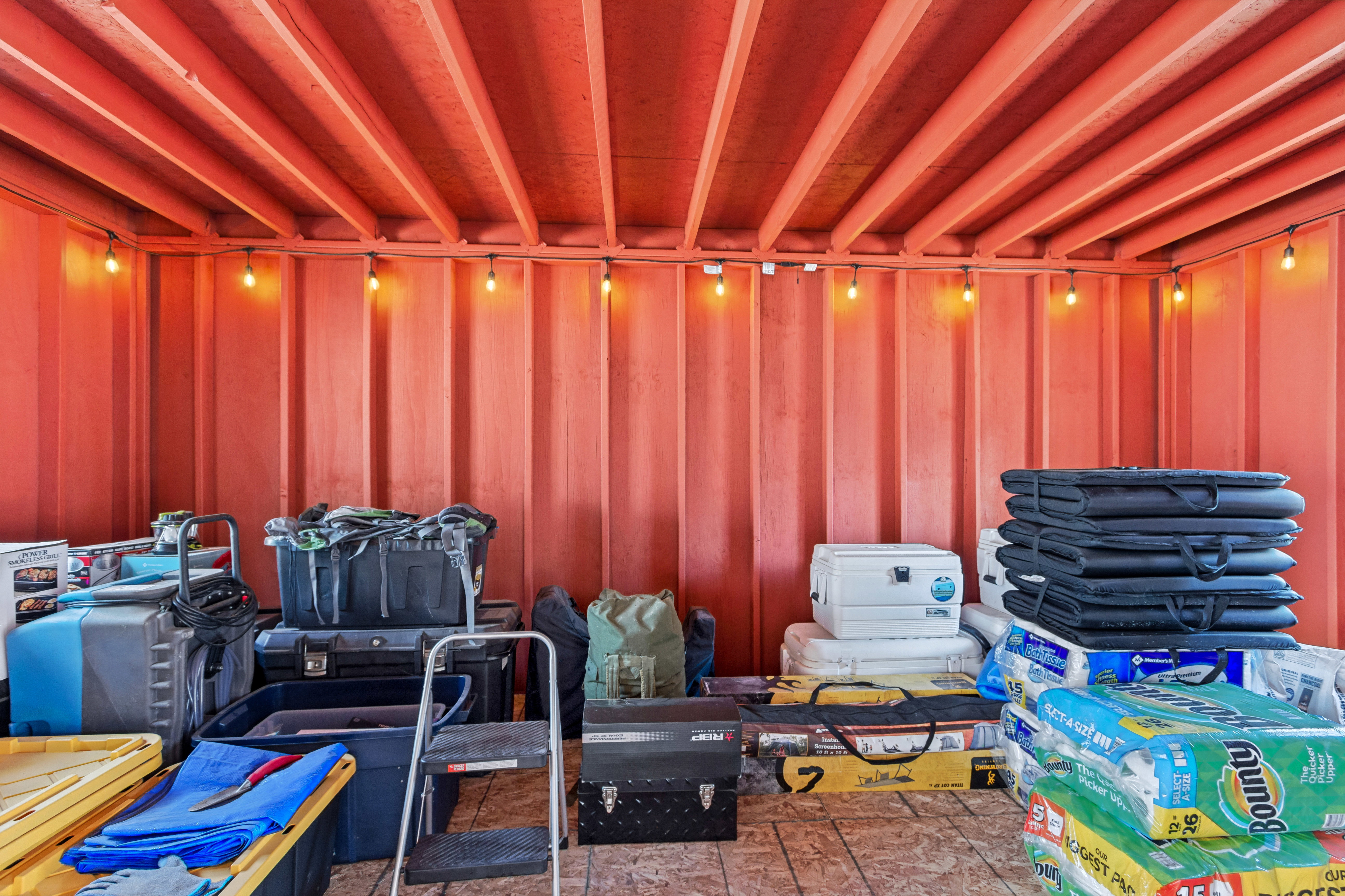 Interior storage space featuring exposed ceiling joists, string lighting, corrugated metal walls, and plywood flooring