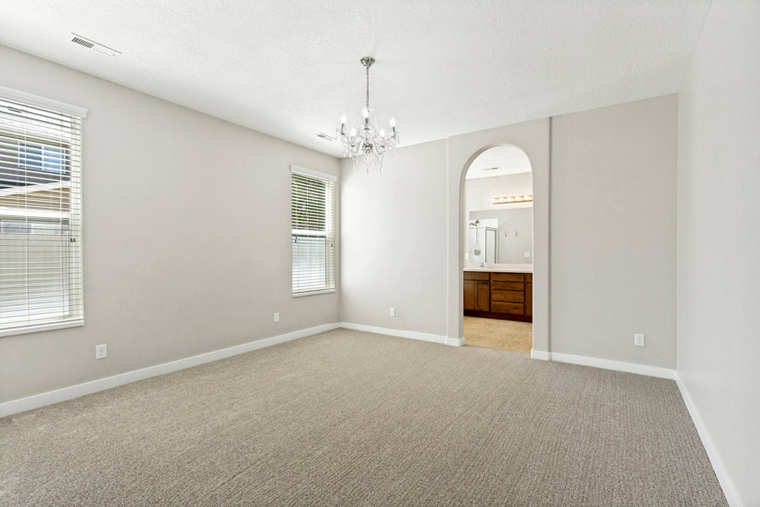 Carpeted primary bedroom featuring a crystal chandelier, two windows with blinds, and an arched doorway opening to a bathroom with a wood-finish vanity and mirror